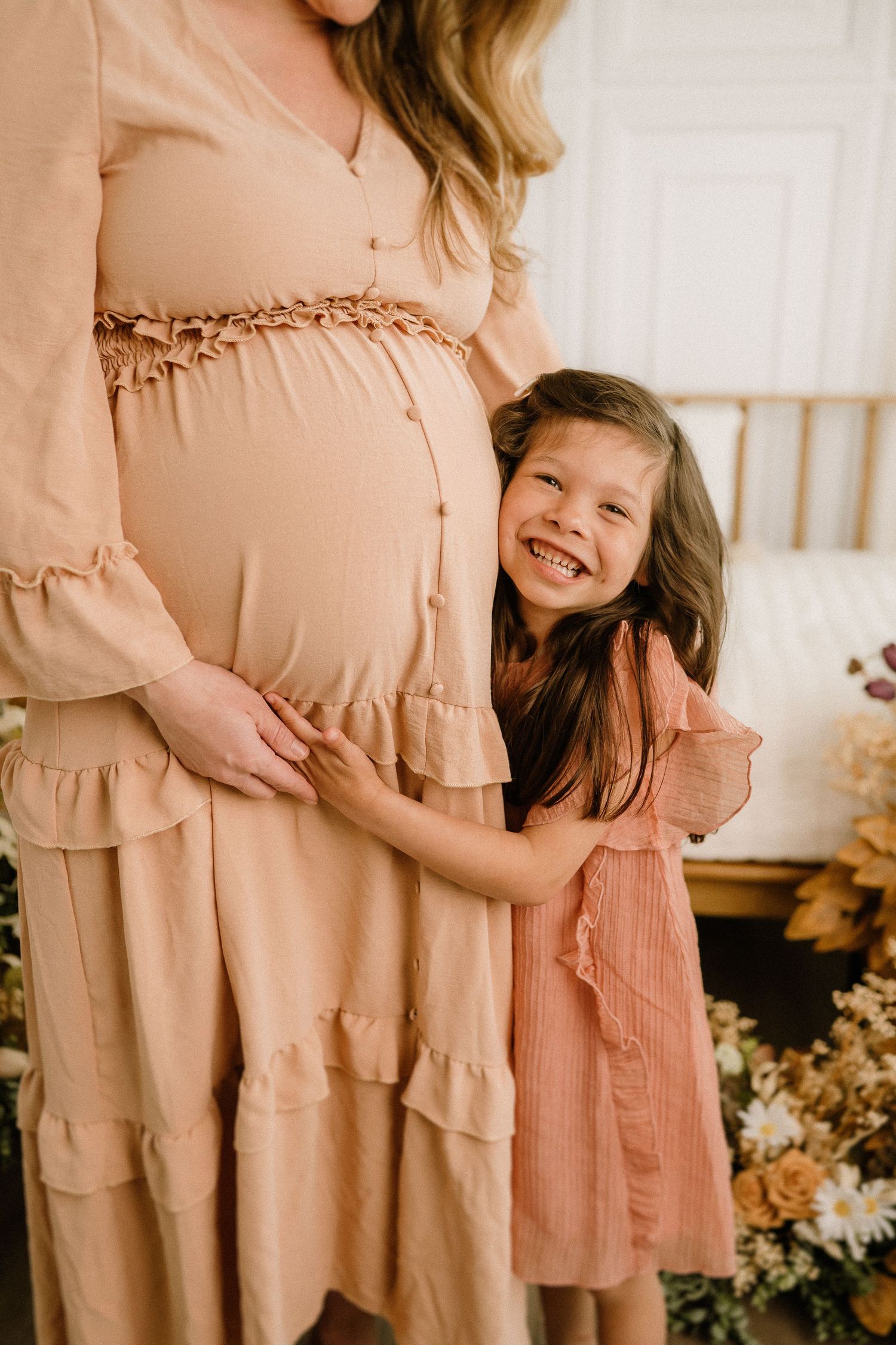 A touching moment with a young child hugging a pregnant woman's belly while wearing matching peach-colored dresses.