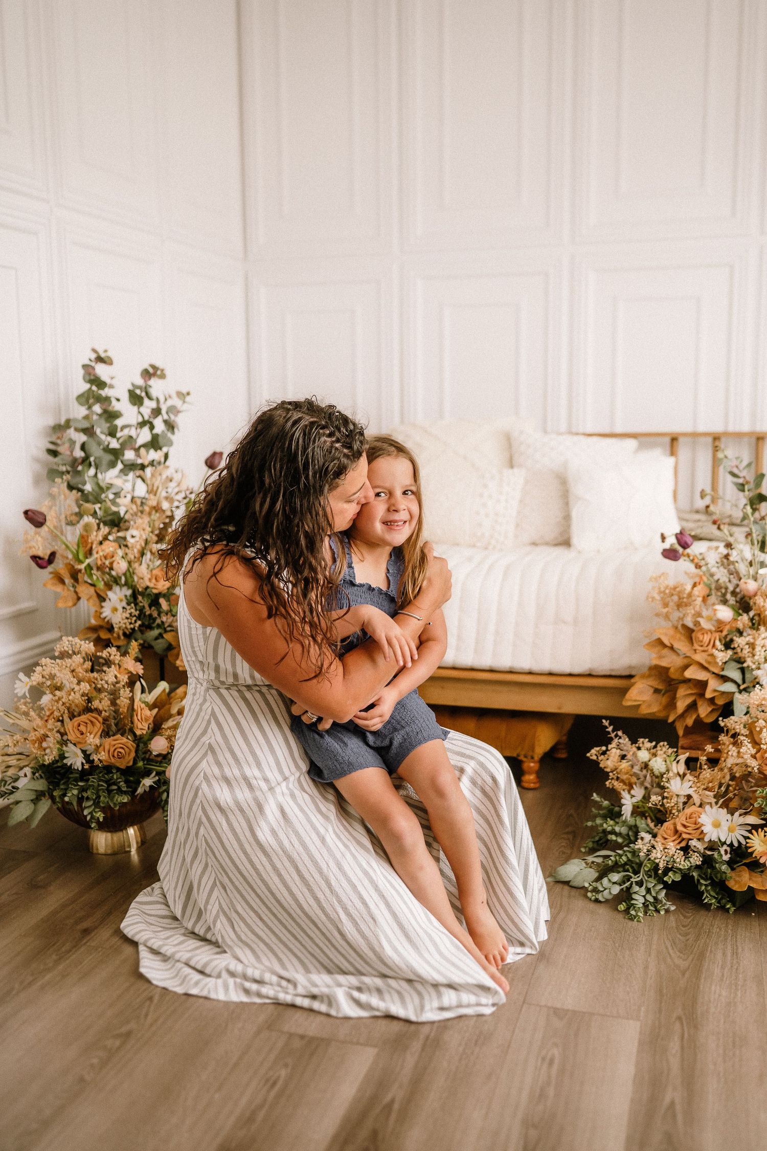 Tender moment between parent and child surrounded by dried floral arrangements in a bright white studio setting.