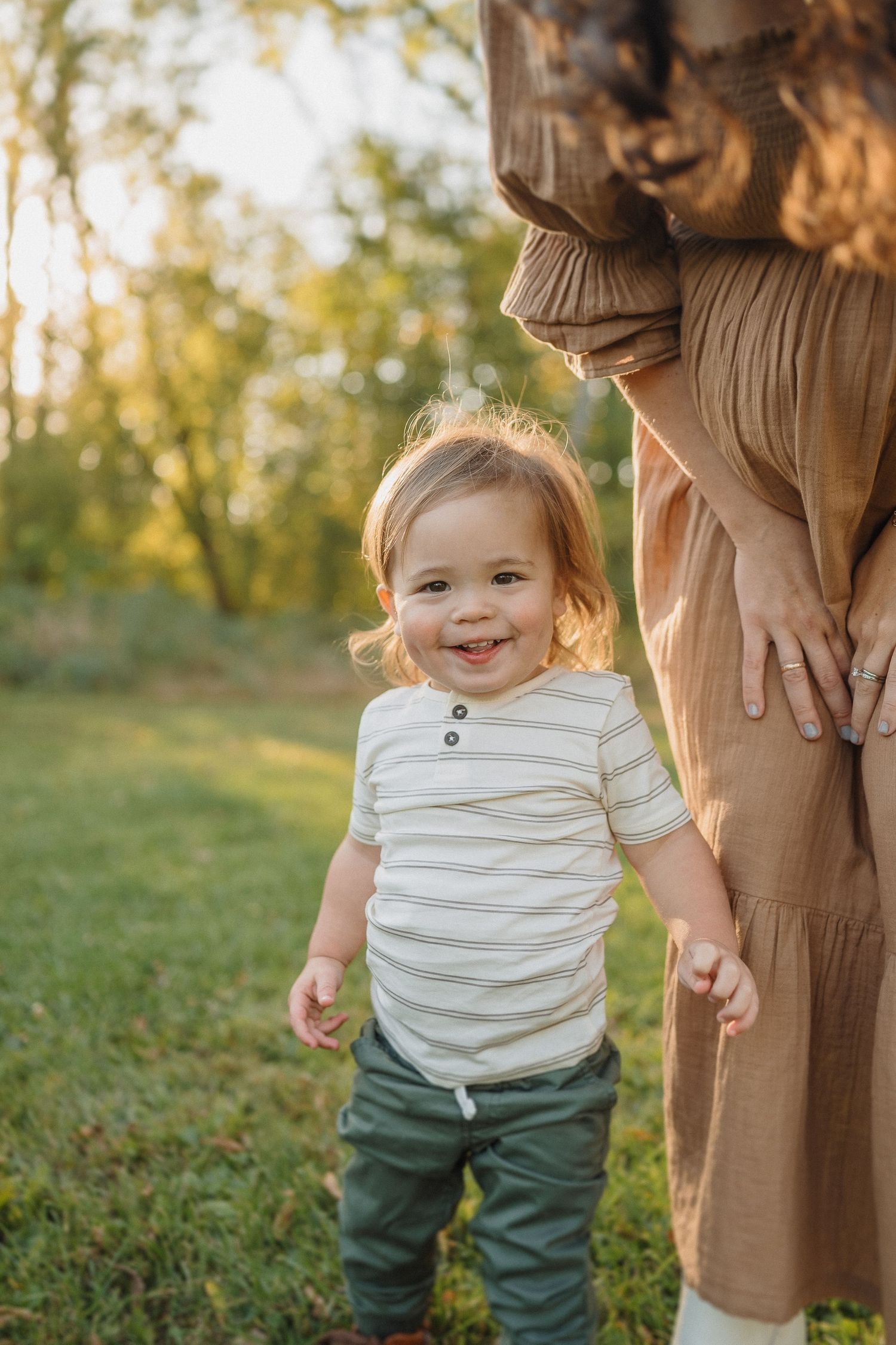 A toddler in a striped shirt smiles while standing next to someone in a brown dress outdoors.