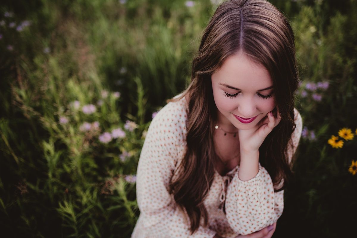 A portrait in a garden wearing a cream polka dot dress with long brown hair looking down while smiling.