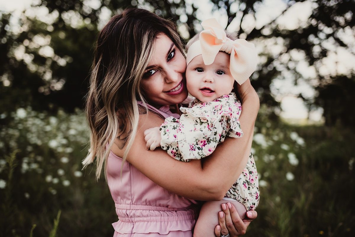Woman in pink dress holding smiling baby in floral outfit outdoors.