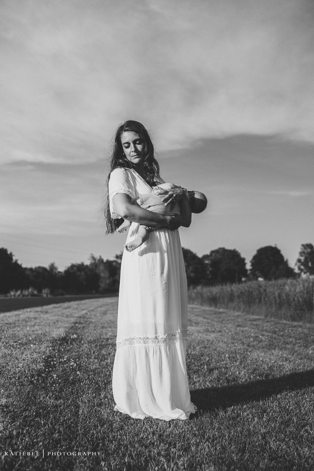 Artistic black and white portrait of woman in flowing white dress standing in field.