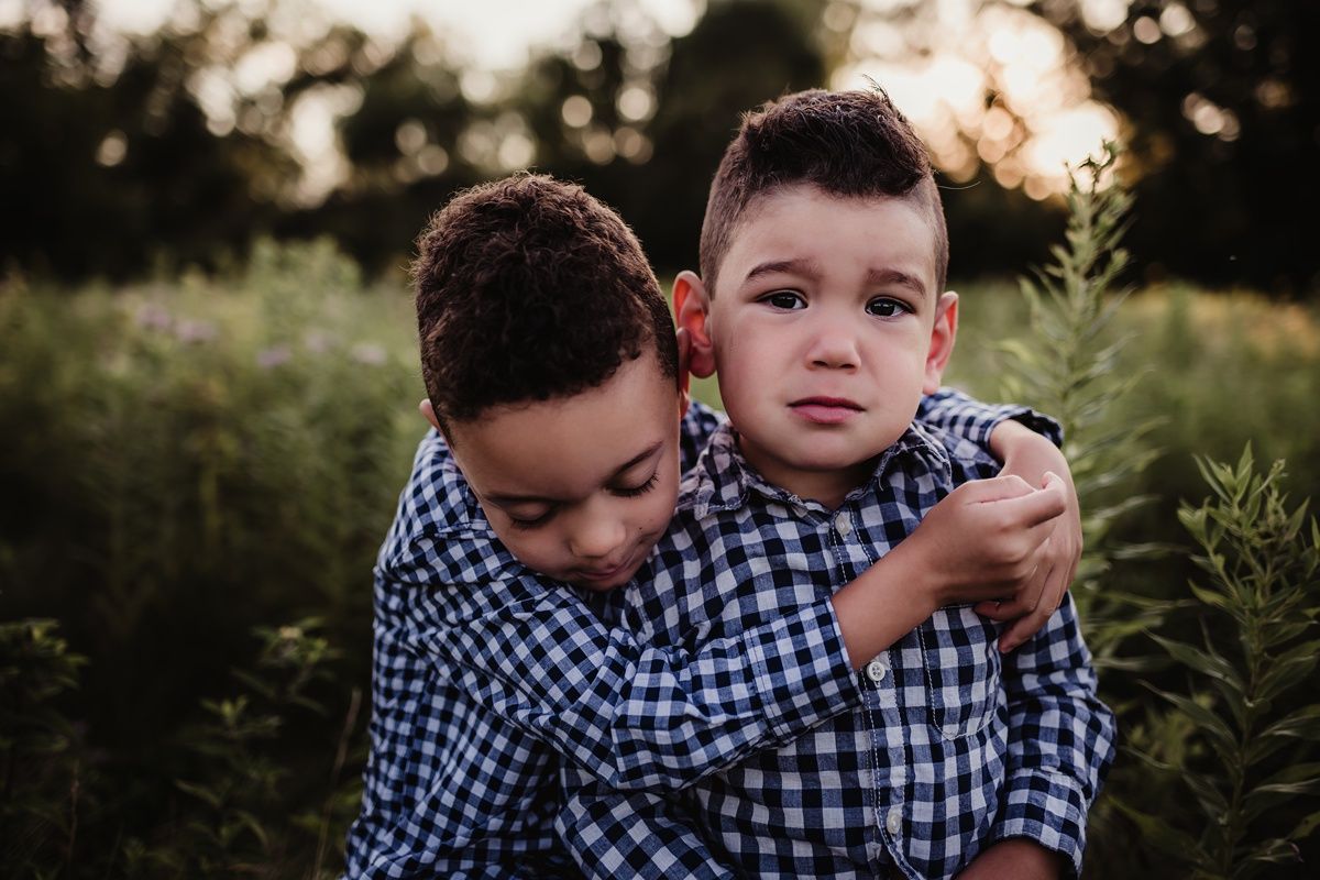 Two young boys in matching blue gingham shirts share an affectionate moment outdoors at sunset.