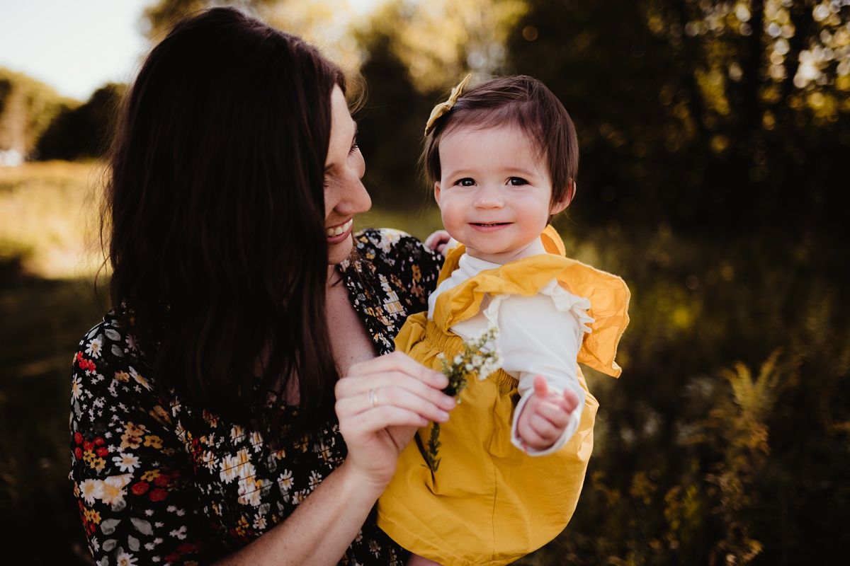 Joyful outdoor portrait with adult in floral dress holding smiling baby in yellow outfit.