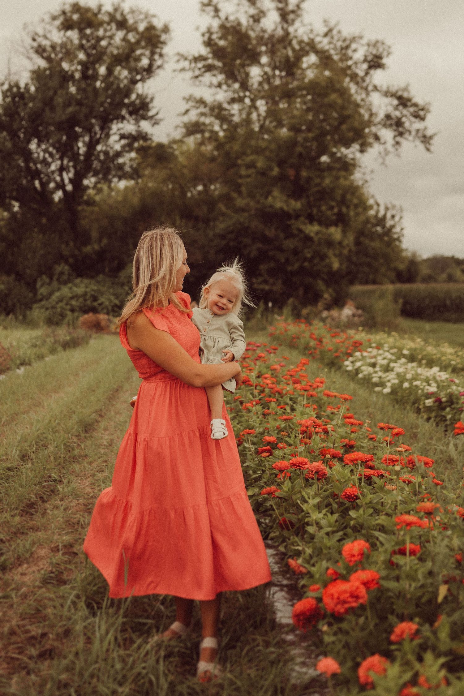 Woman in coral dress sharing a tender moment with child among blooming red zinnias.