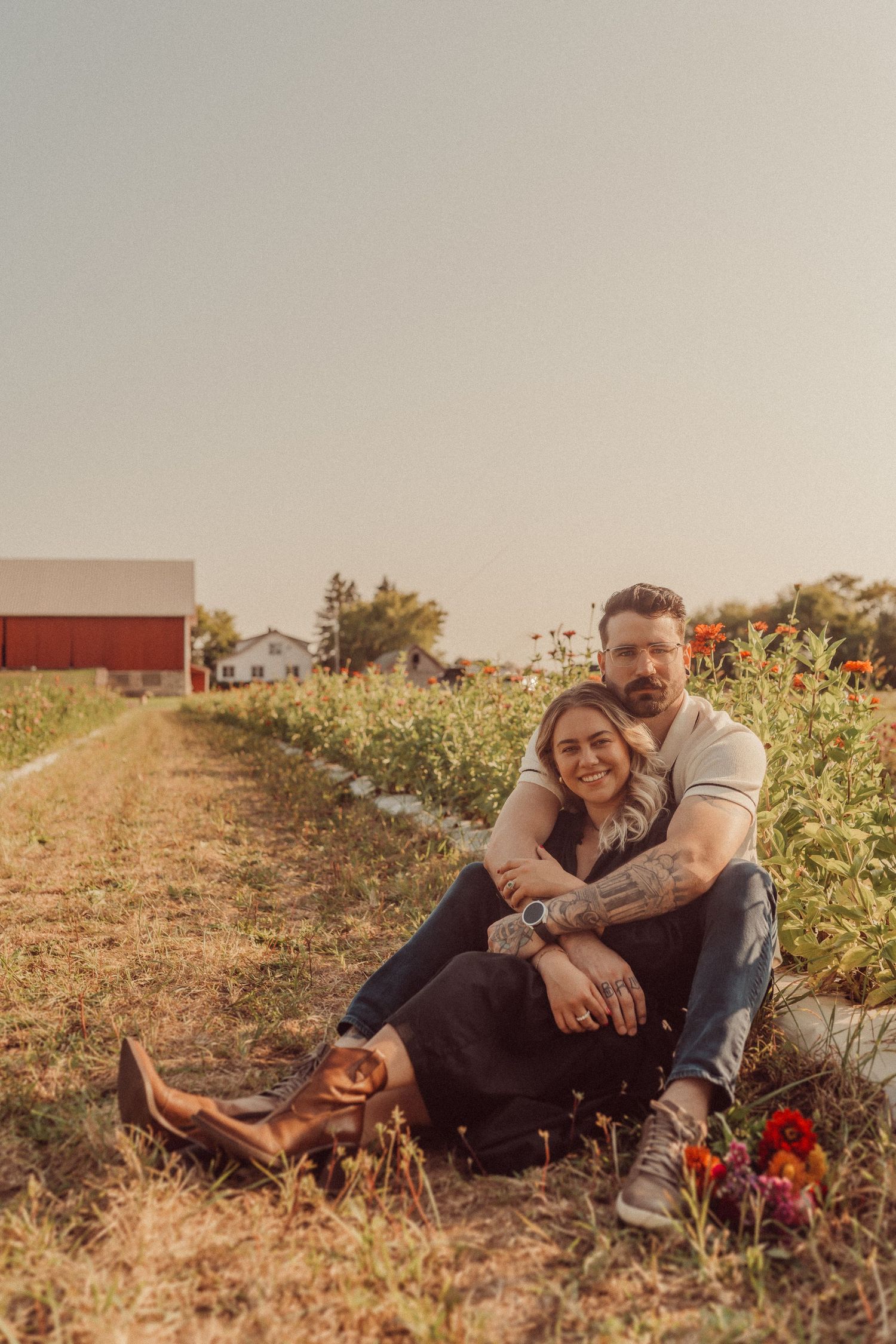 Couple relaxing together in a flower field with a red barn in the background.