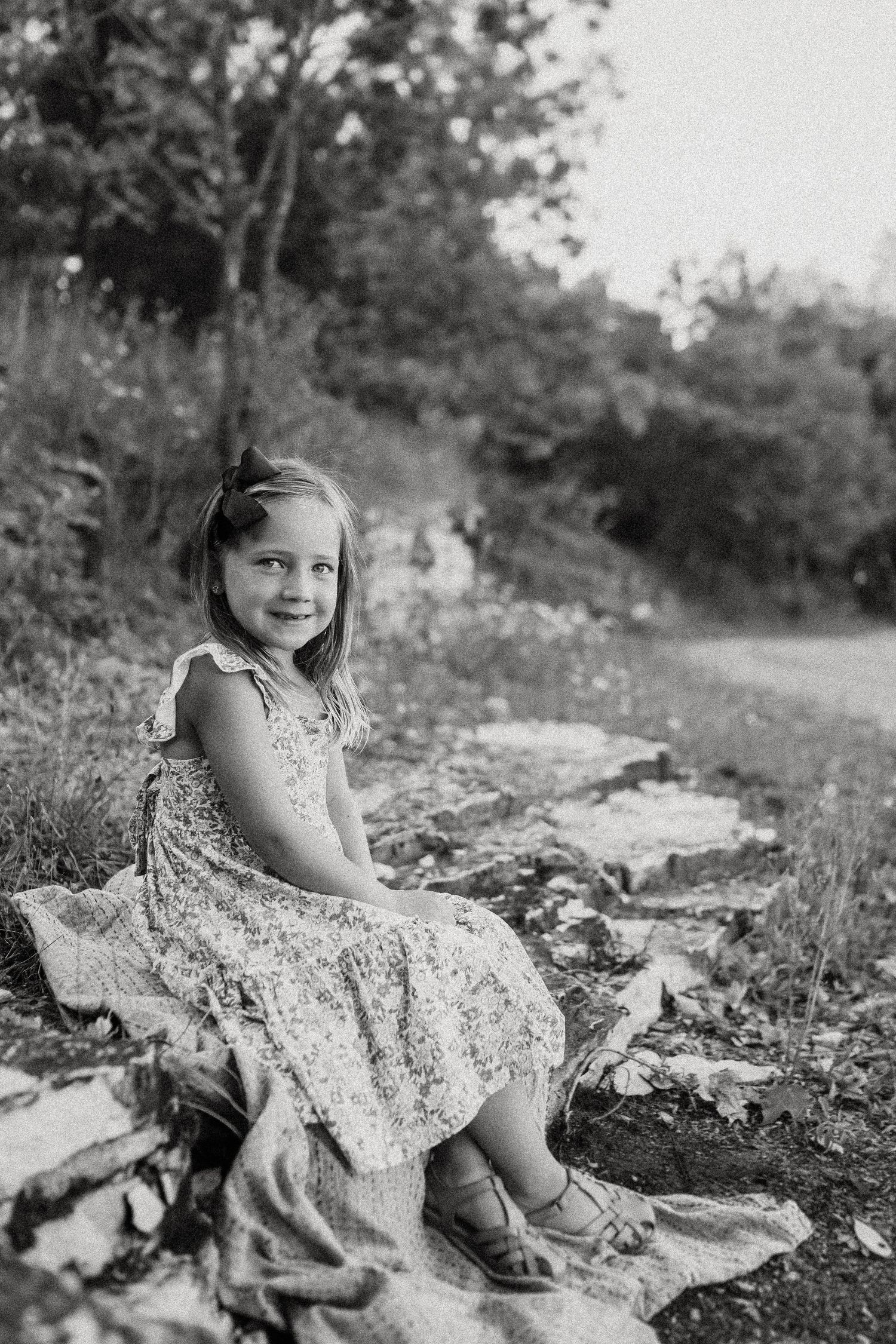 A black and white portrait of a child sitting on rocky ground in a floral dress.