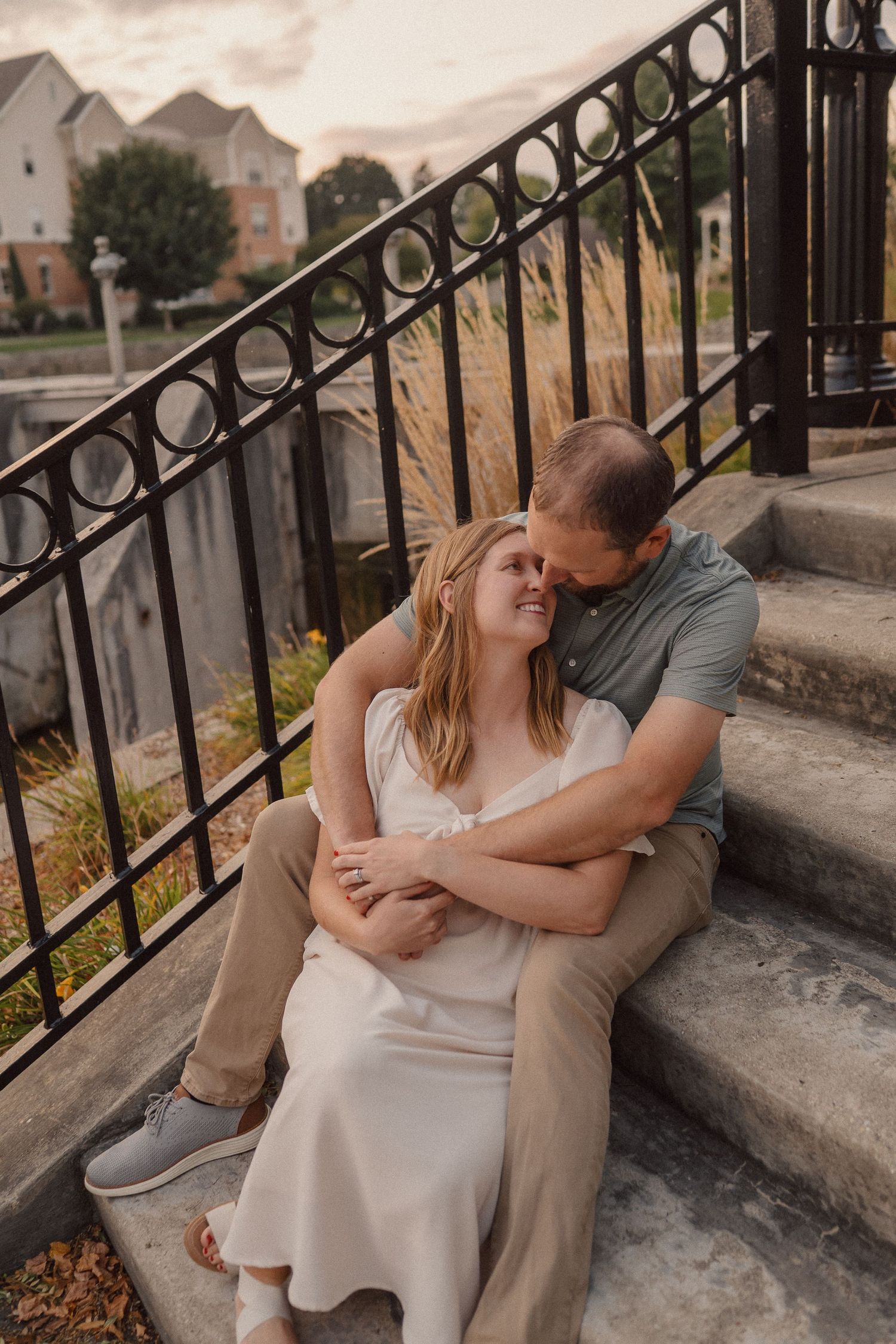 A romantic couple sits on stone steps with an ornate metal railing, sharing an intimate embrace at sunset.
