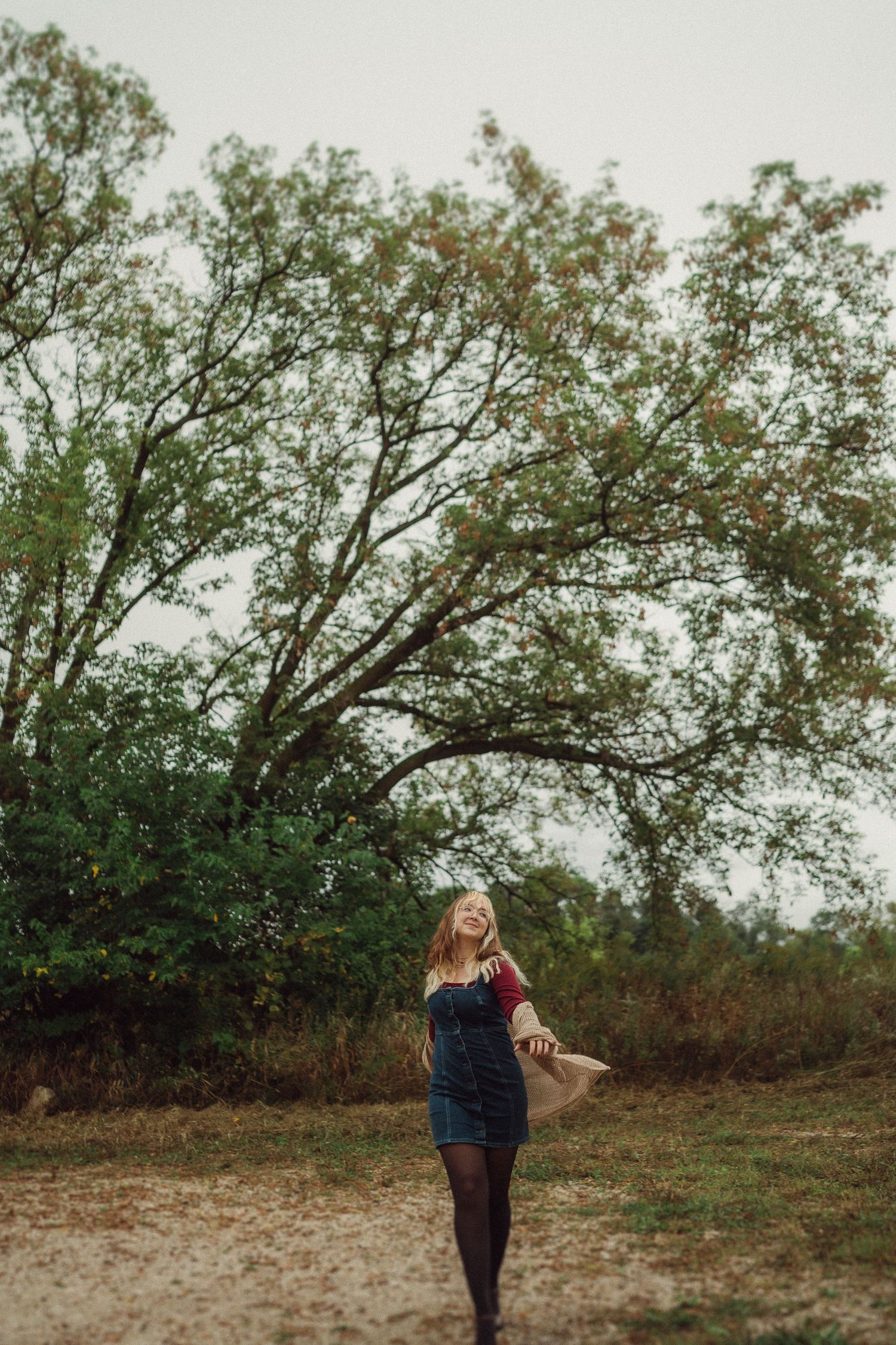 Stylish figure in denim dress poses beneath autumn trees on dirt path.