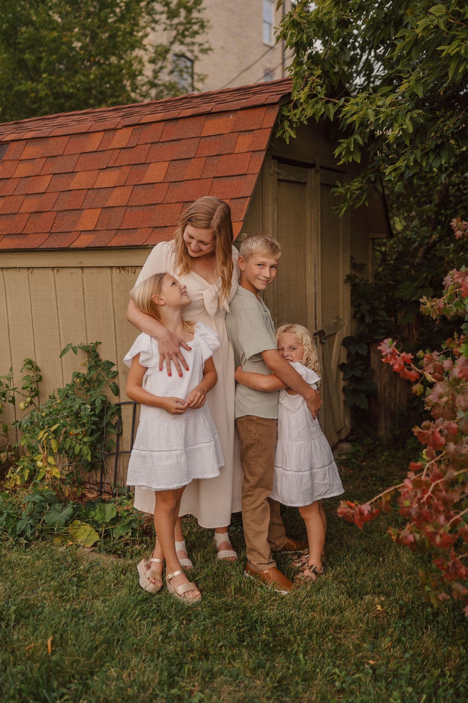 Family group poses playfully near a garden shed with red roof and flowering plants.