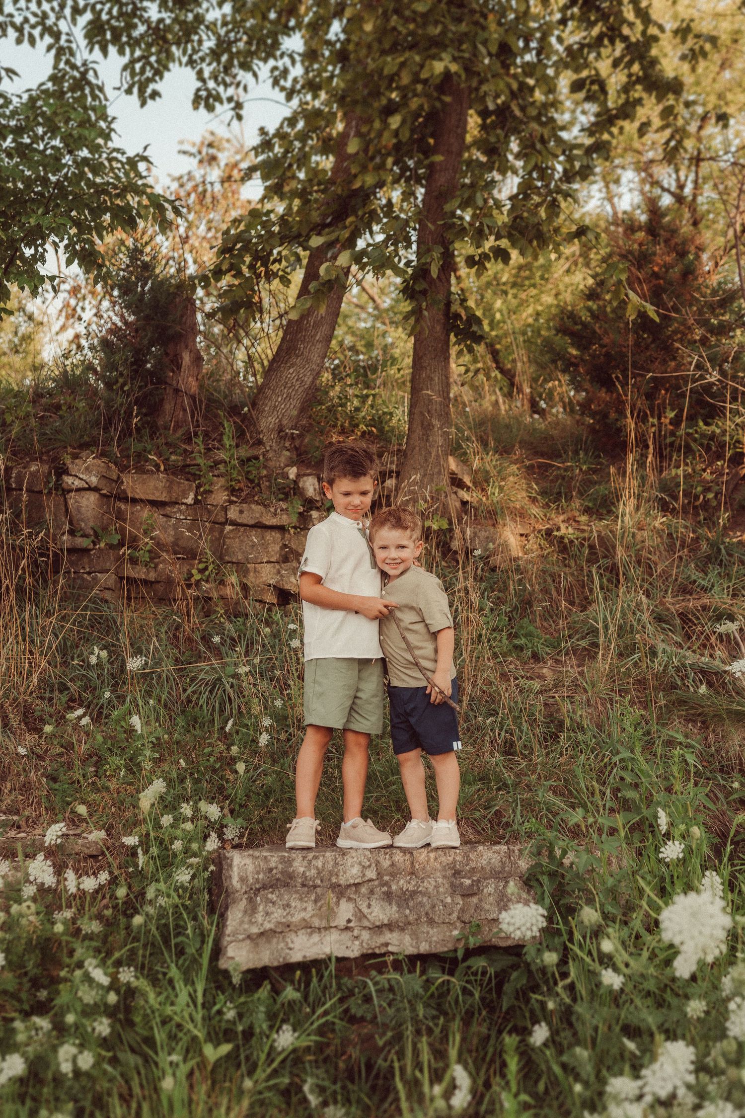 Two children stand together on a stone wall in a wooded area during golden hour.