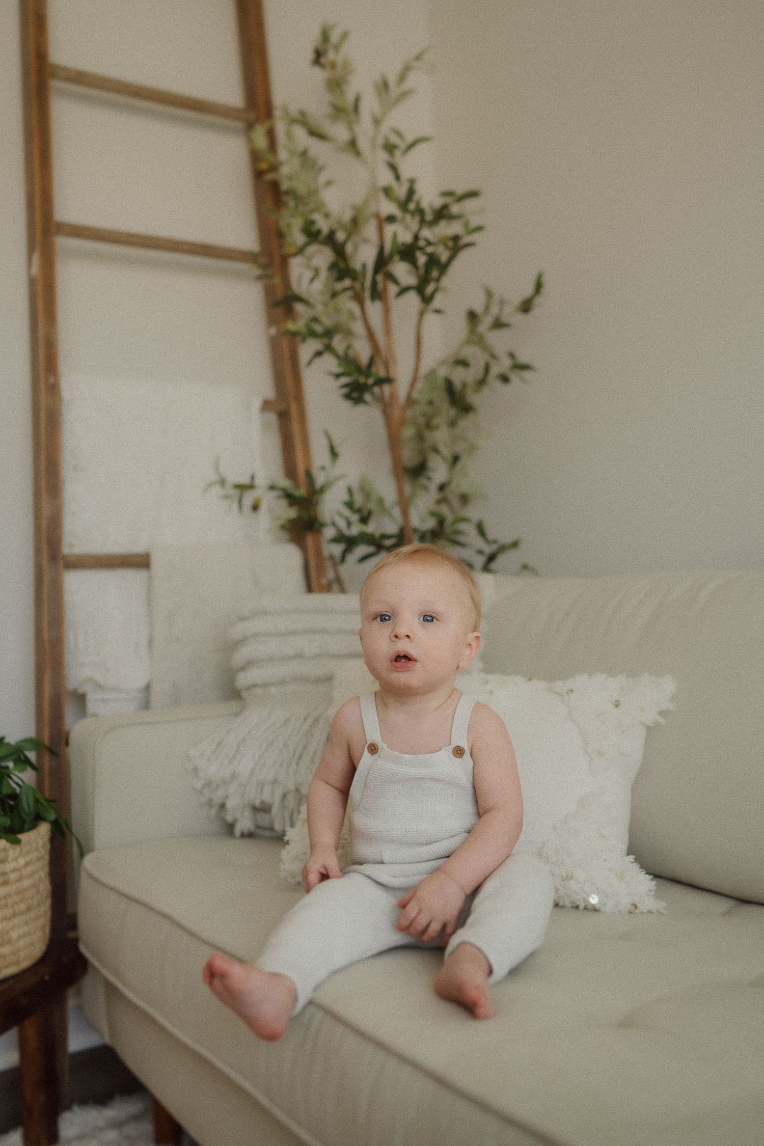 Baby in white outfit sits on beige couch with decorative ladder and greenery in background.
