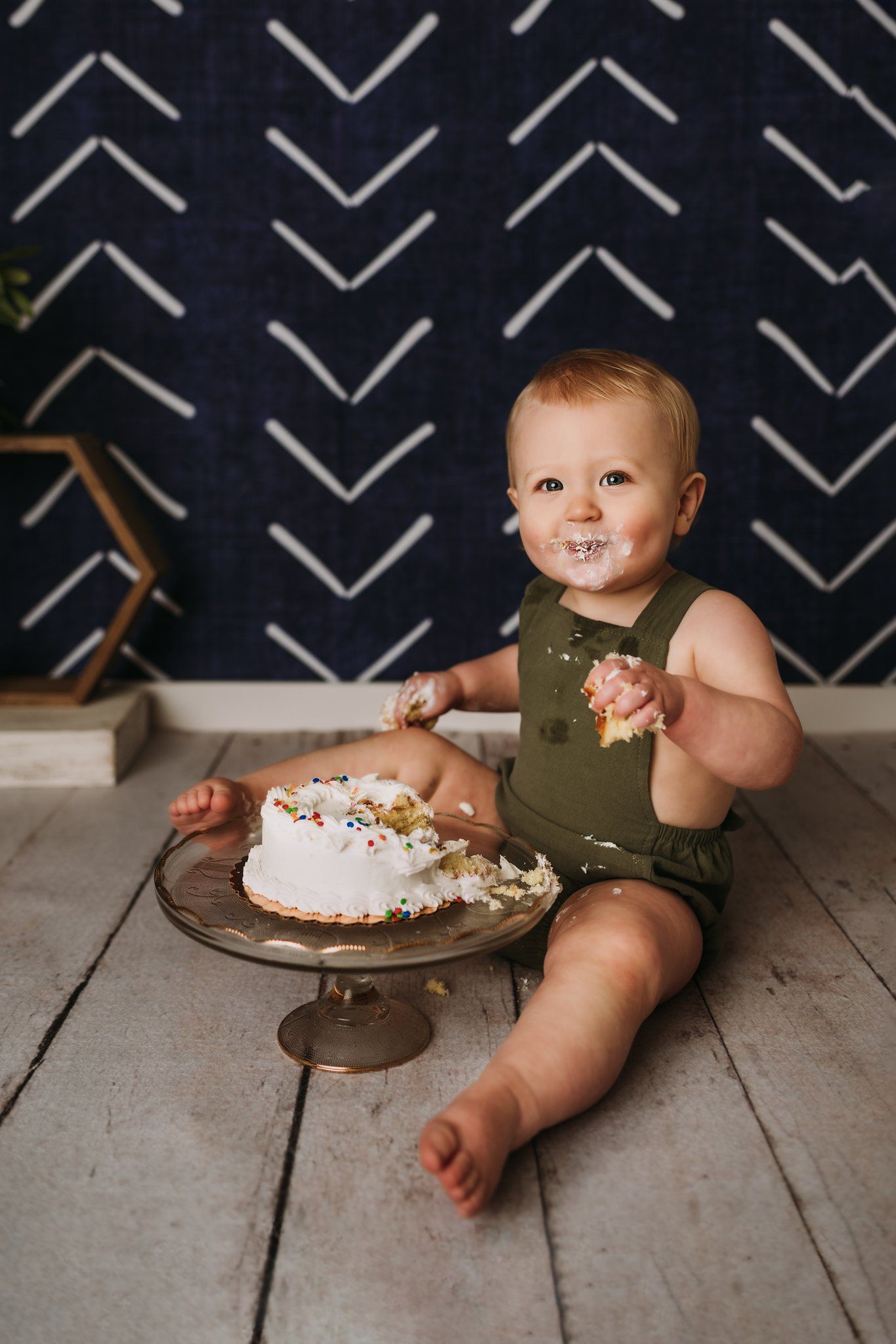 A cake smash photography session with a white frosted cake on a pedestal against a navy chevron wall.