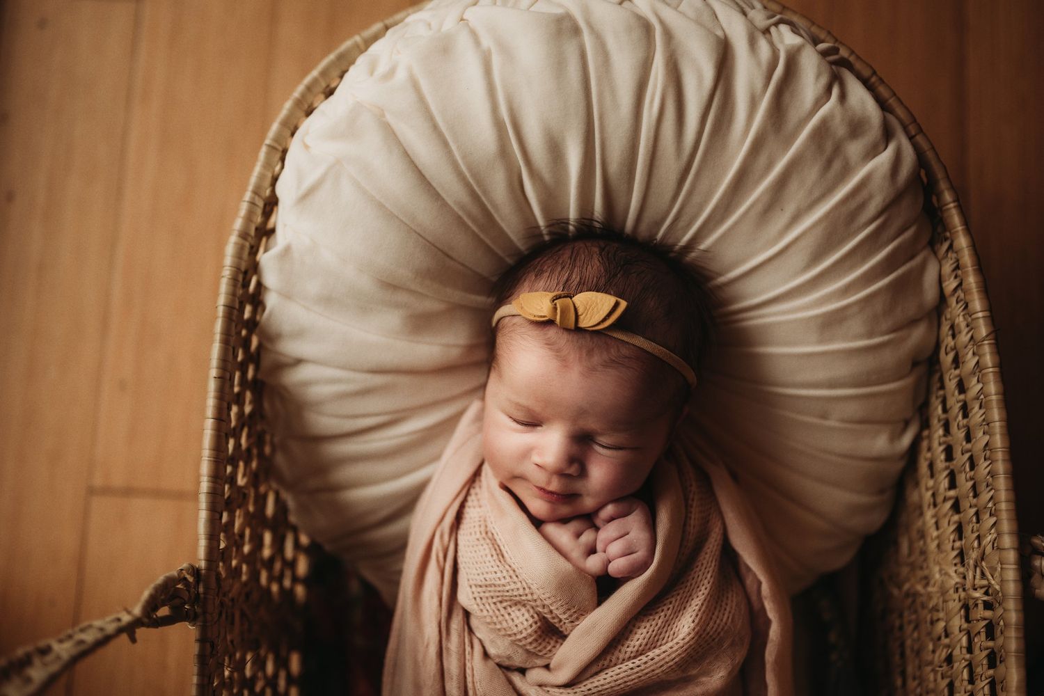 Sleeping newborn wrapped in beige fabric resting on circular cushion with delicate bow headband.