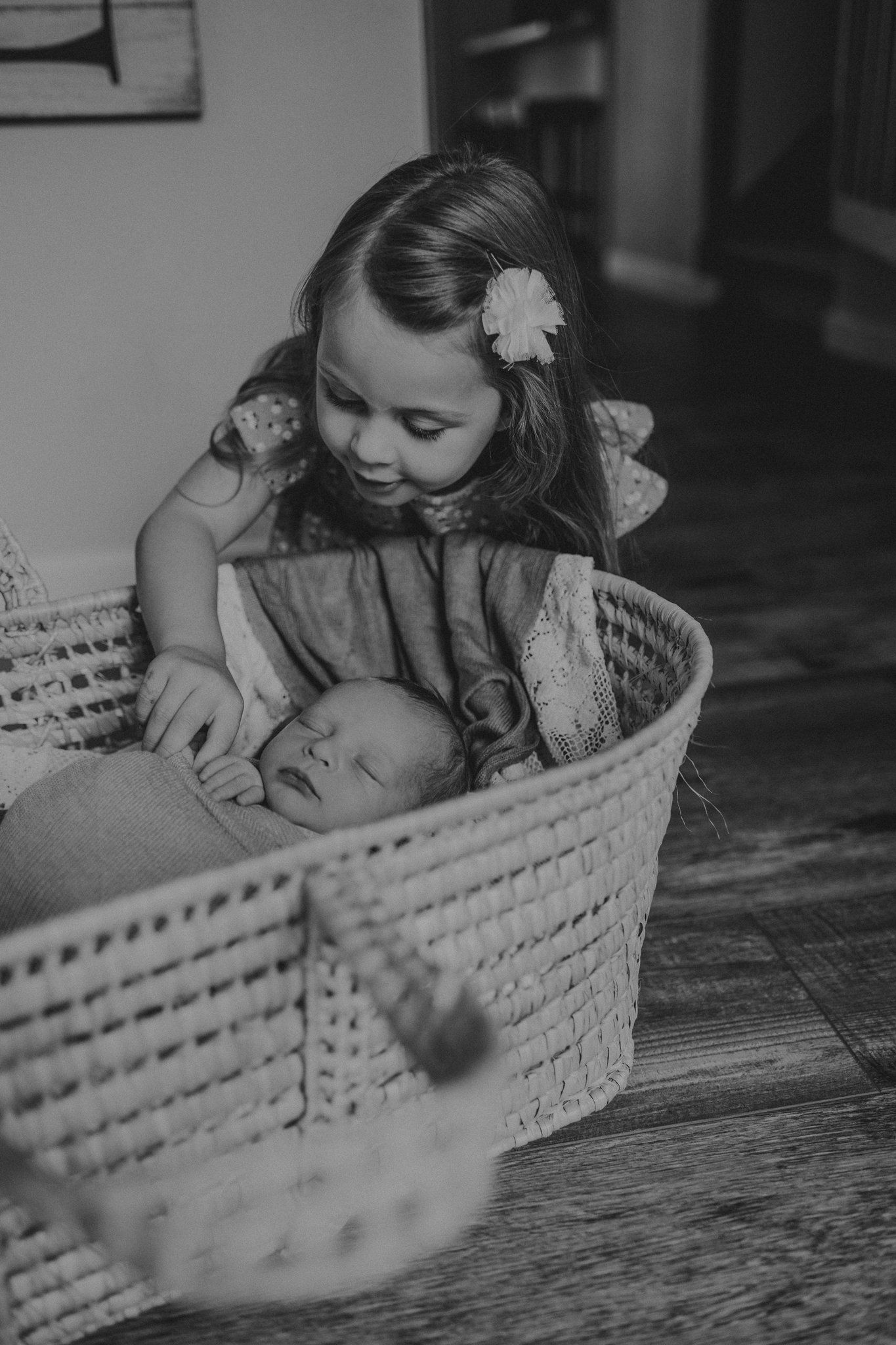 Black and white image of child looking down at infant resting in woven basket.