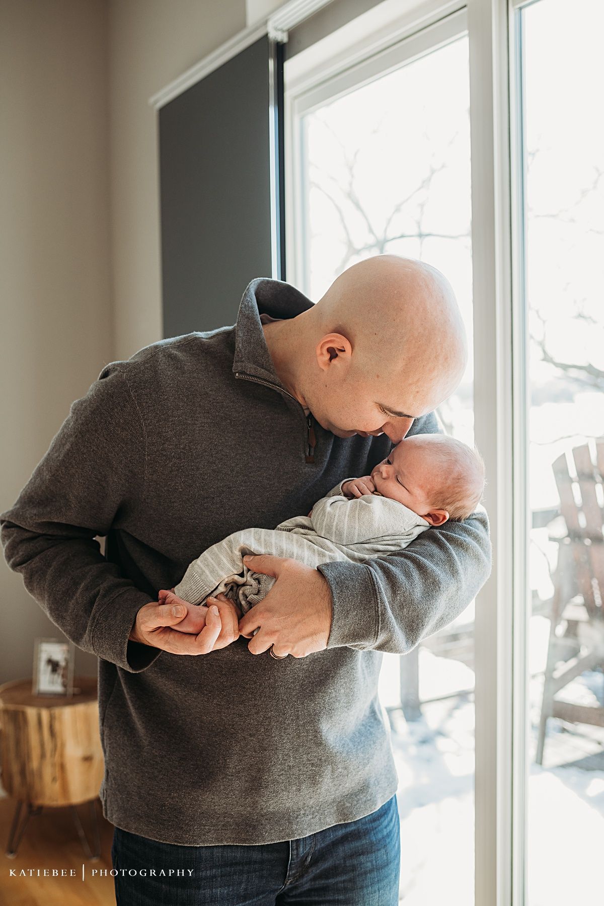 Parent in gray sweater tenderly holds and kisses sleeping newborn by bright window.