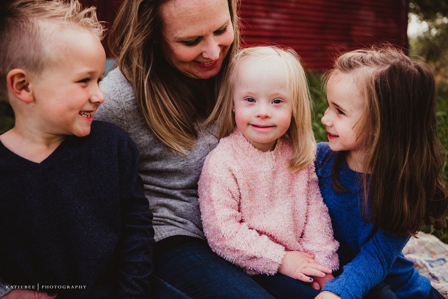 Family sits close together sharing smiles against red barn backdrop.
