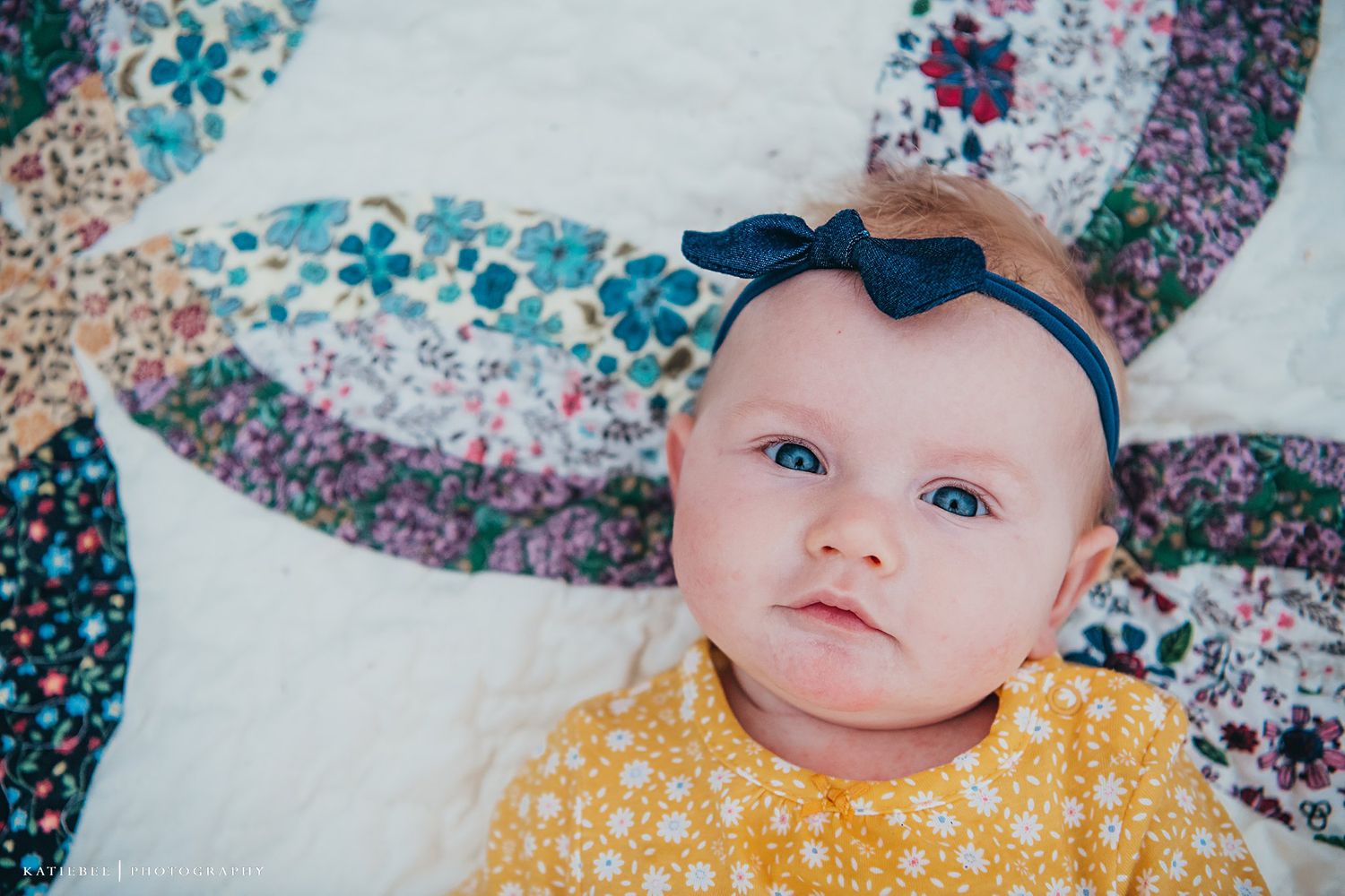 Baby wearing yellow polka dot outfit and navy bow on colorful patchwork quilt.