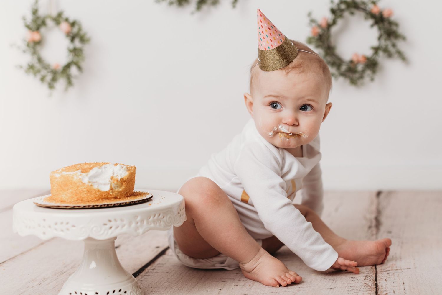 Baby in party hat sitting beside cake on white backdrop with decorative wreaths.