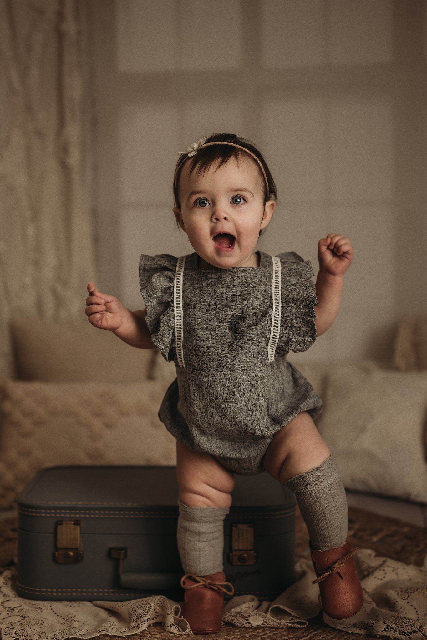 Baby in grey ruffled romper stands energetically on vintage suitcase.