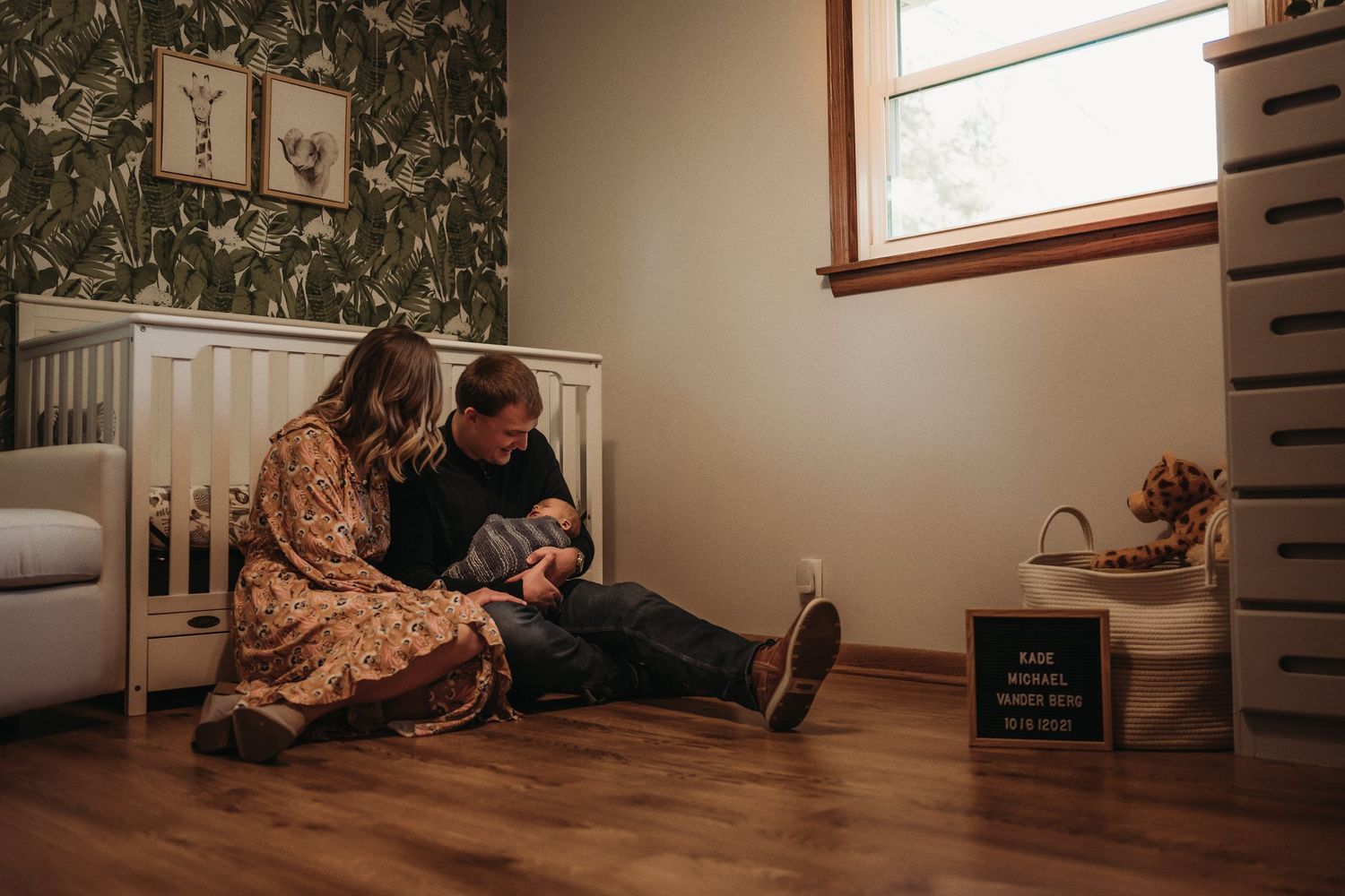 Cozy nursery scene with floral wallpaper and white crib as couple sits on hardwood floor.