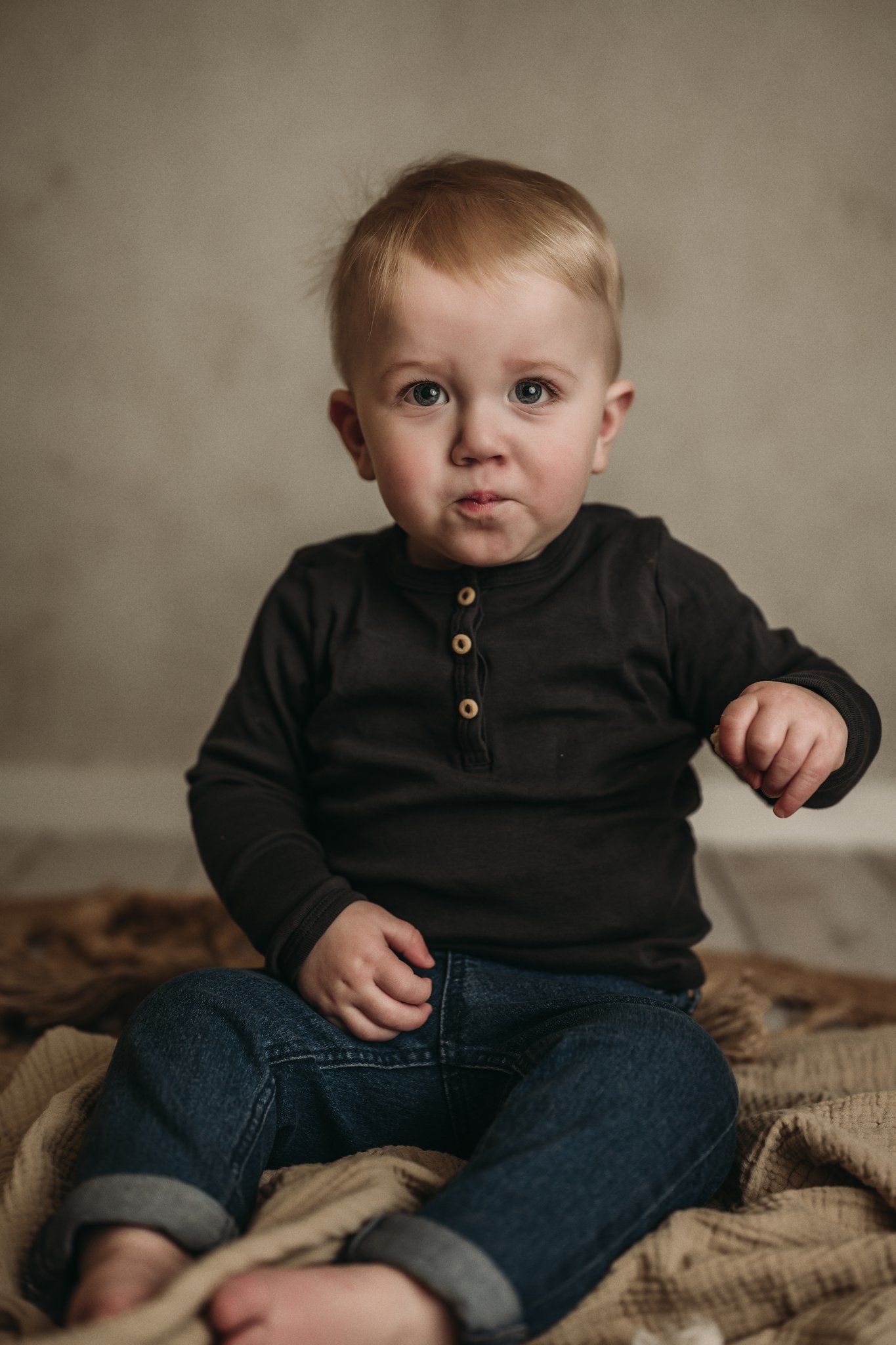 Indoor portrait of a toddler in black henley and rolled jeans sitting on a textured blanket.