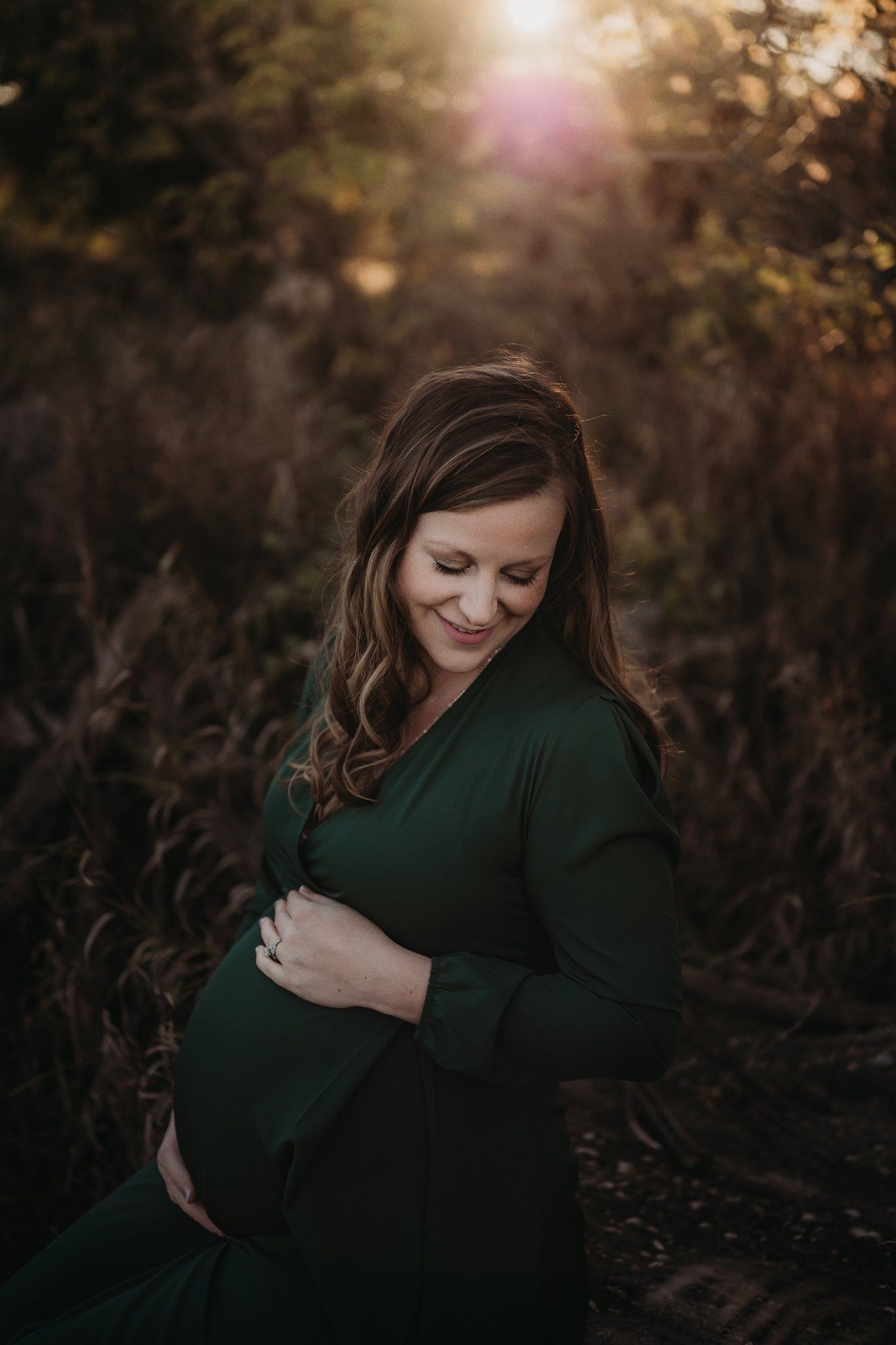 A maternity portrait in a forest with golden sunlight filtering through trees.