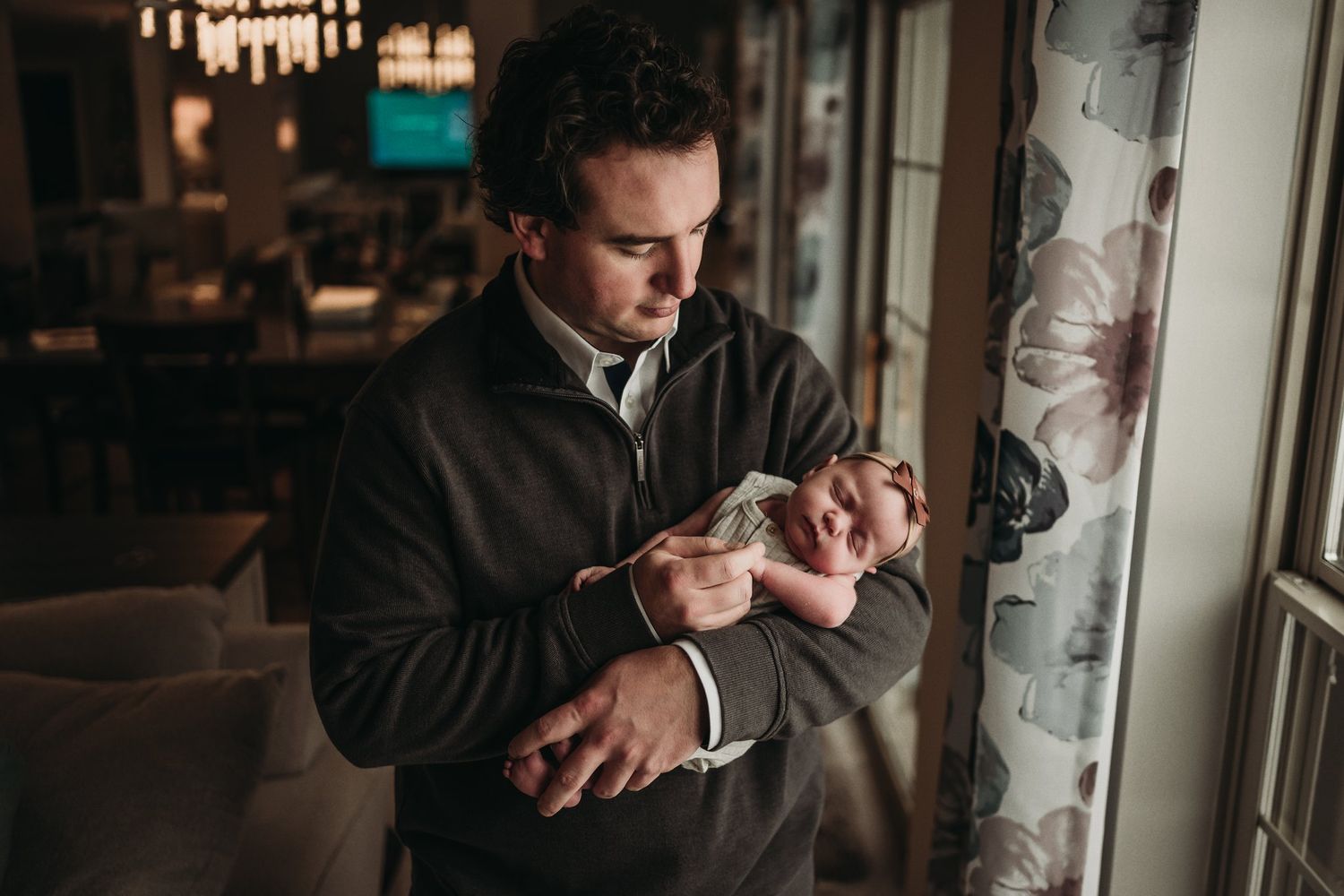 A parent wearing a dark sweater gazes down lovingly at their newborn by a window.