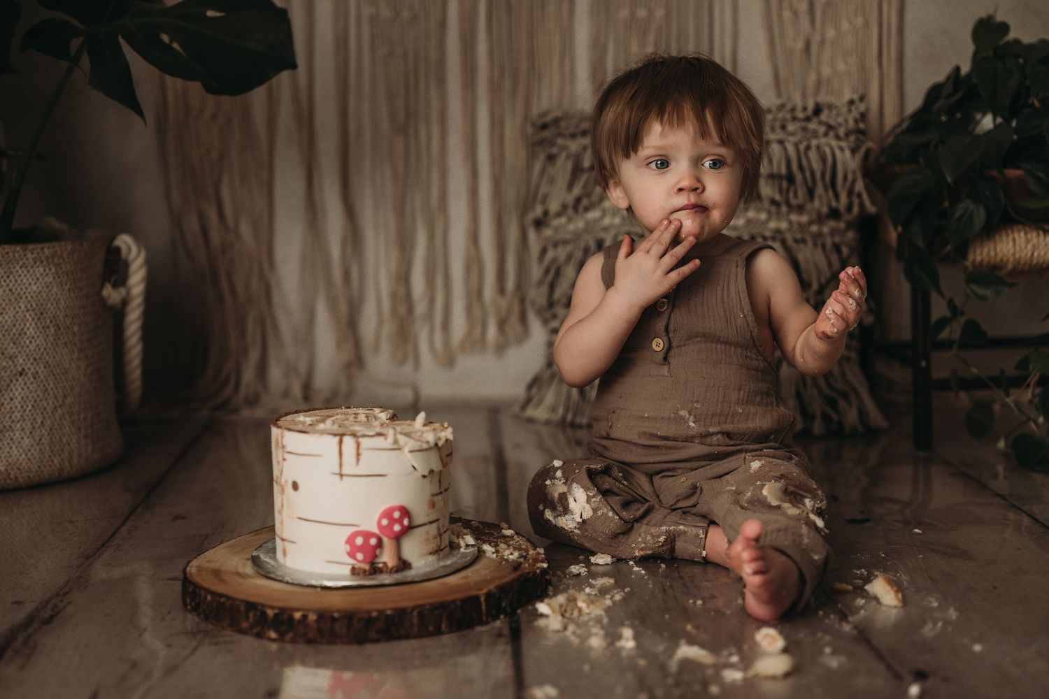 Toddler in brown romper eating cake during messy first birthday cake smash session.