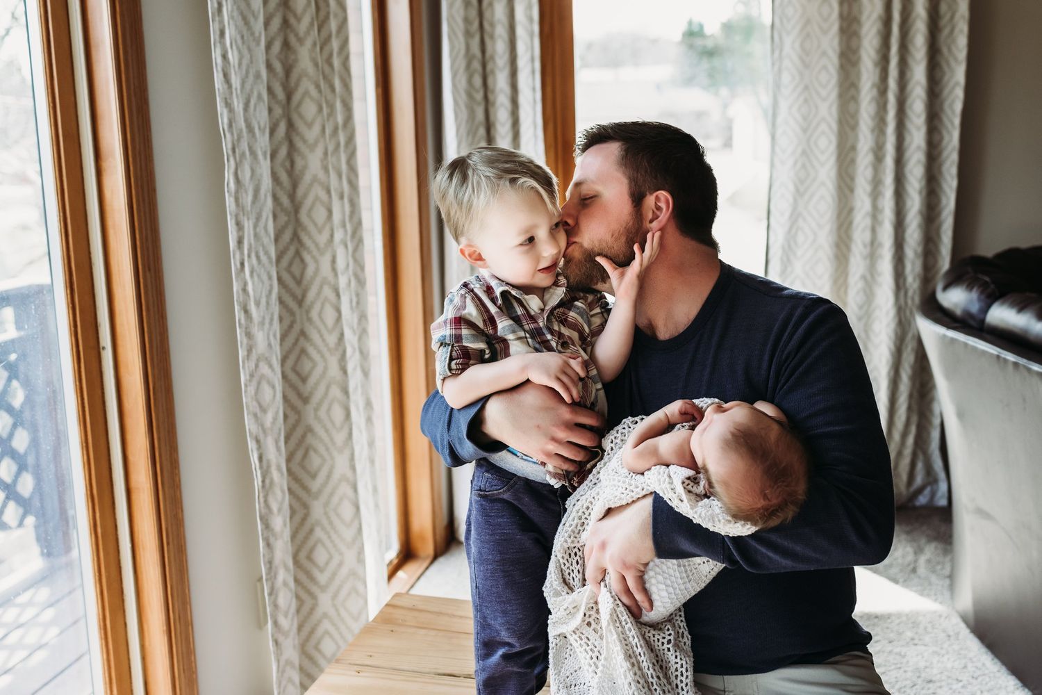 Family shares a sweet moment by large windows as parent juggles holding both children.