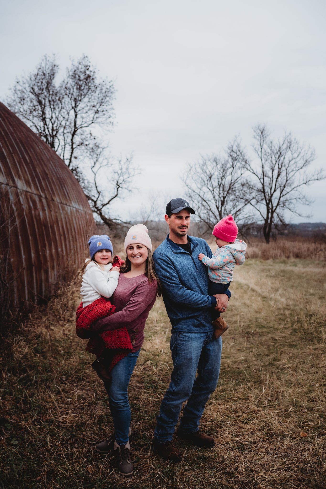 Full length family portrait in casual winter wear against rustic curved barn wall.