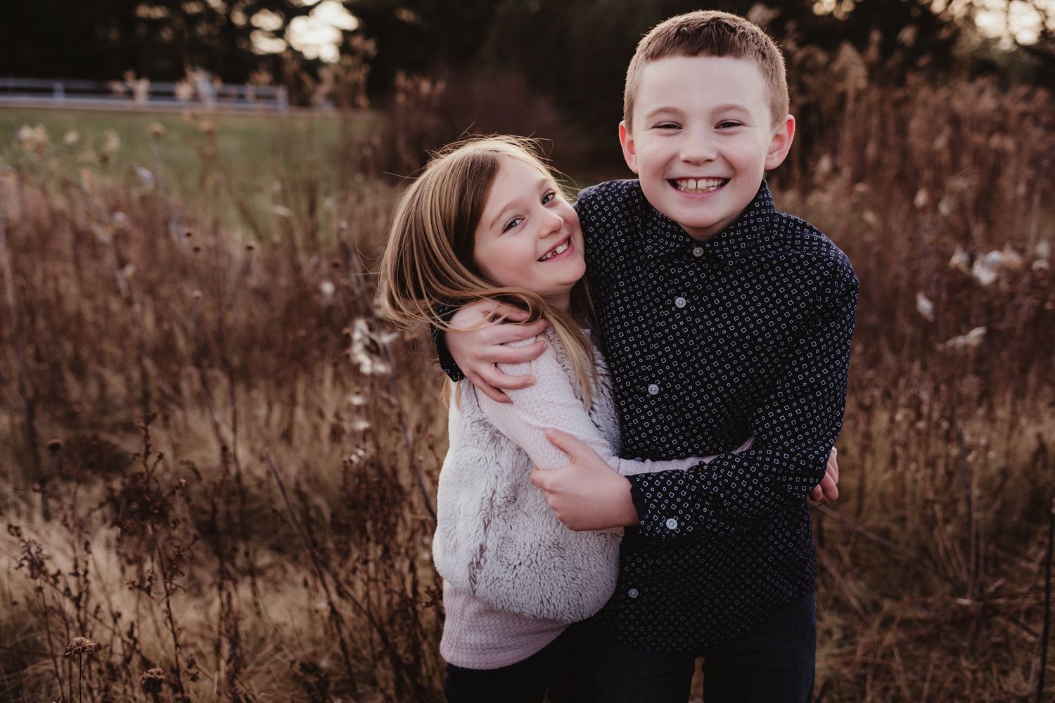 Two children hugging and laughing together in a field of tall grass at sunset.