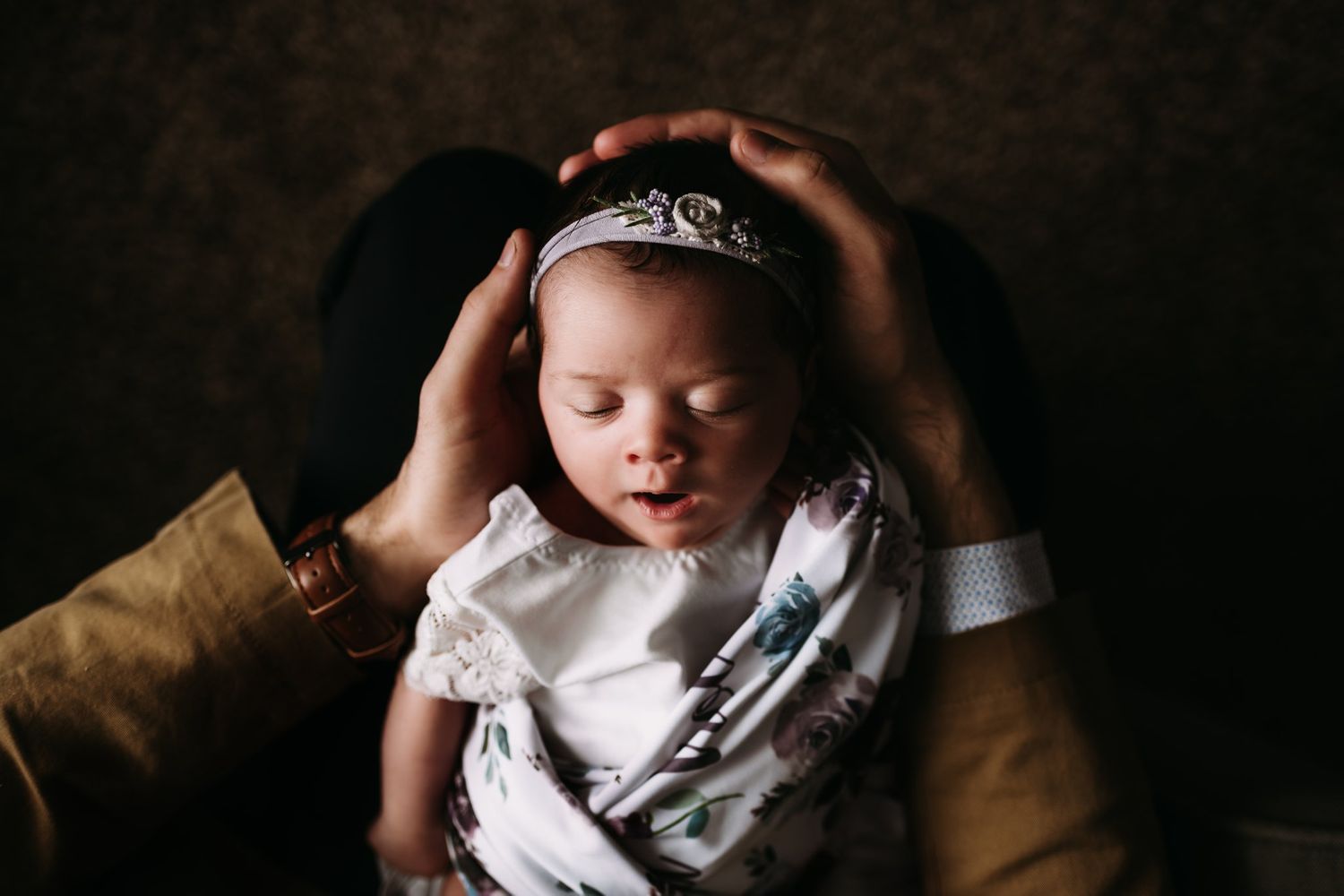 A baby wearing a delicate white dress and tiara rests peacefully on a brown chair.