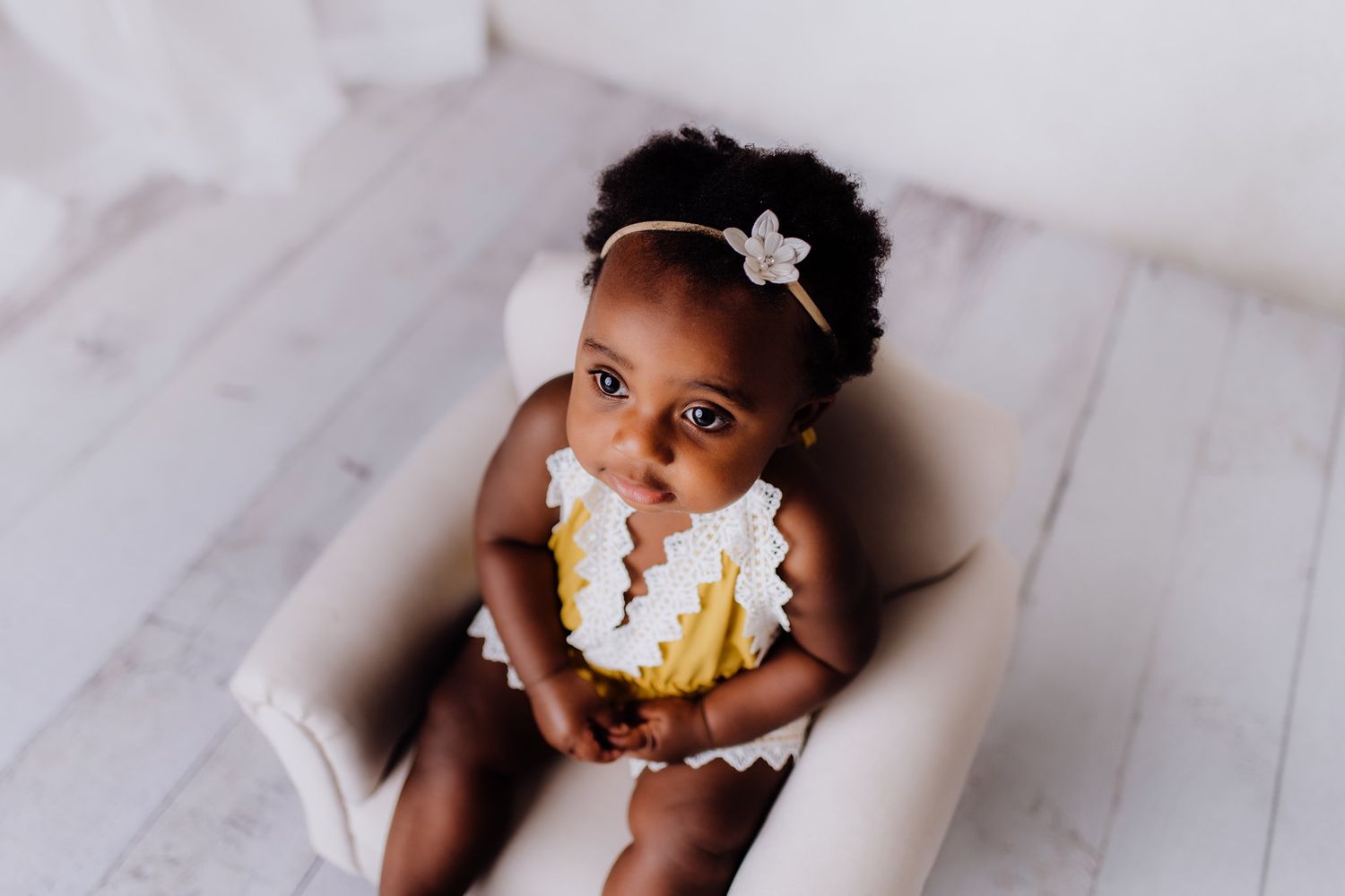 A small child wearing a ruffled outfit sits in a beige chair with a happy expression.