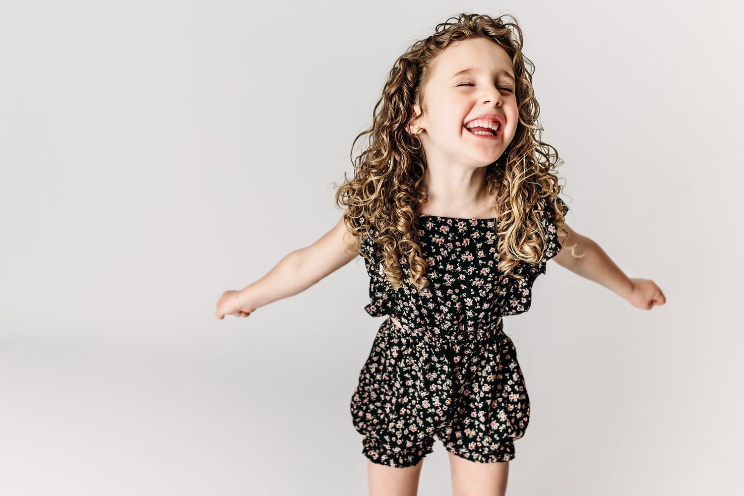 A joyful child wearing a black floral romper dances against a white backdrop.