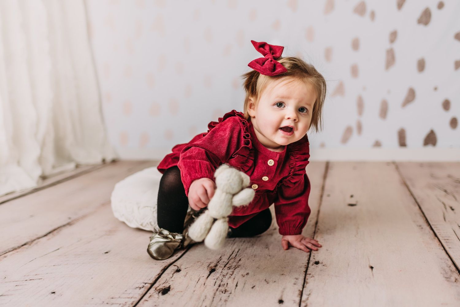 Baby in red ruffled dress and bow plays with stuffed toy on rustic floor.