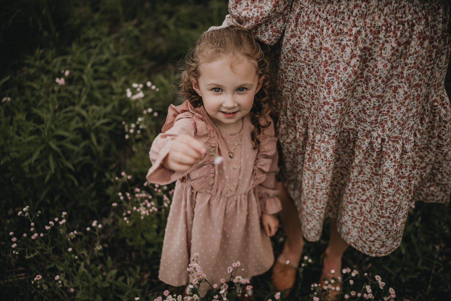 Young child in ruffled dress stands among wildflowers in a natural outdoor setting.