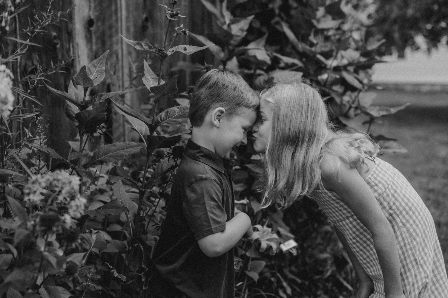 Black and white portrait of two children sharing a sweet moment by garden wall.