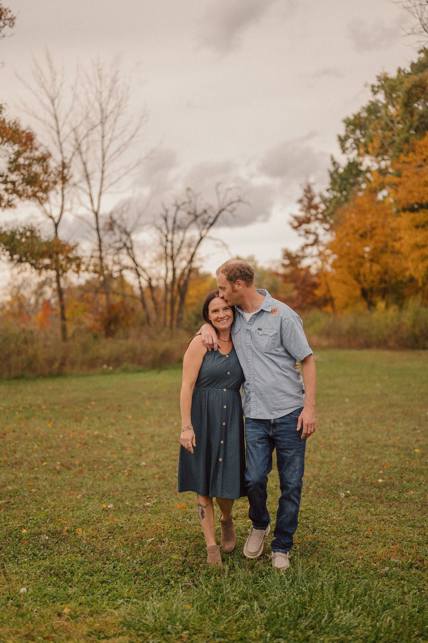 A couple enjoys an intimate moment in a grassy field with autumn trees in the background.
