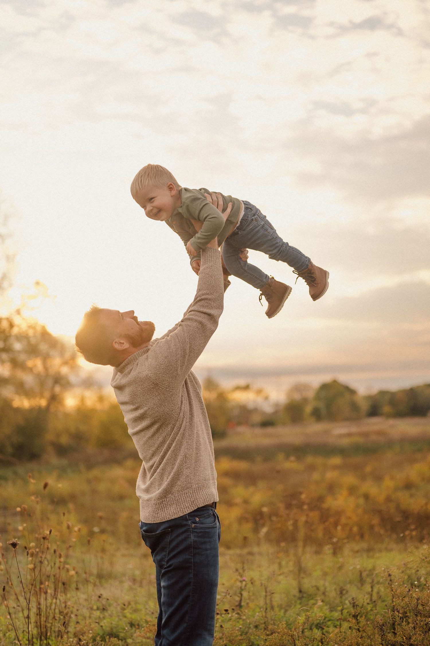A parent in a gray sweater lifts and plays with a small child in a golden sunset-lit meadow.