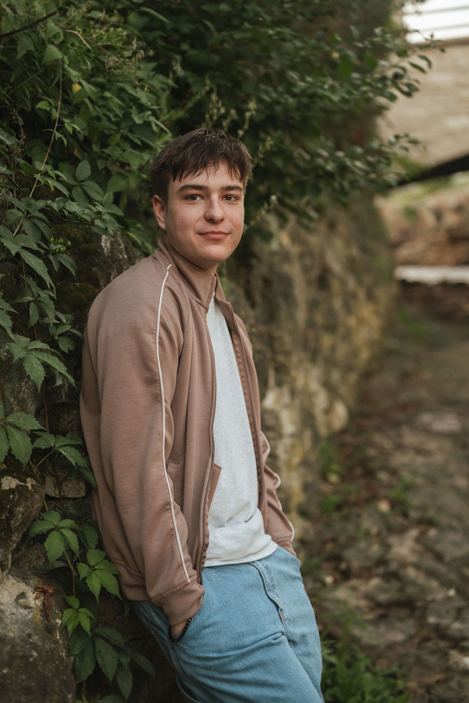 A casual outdoor portrait against a leafy background wearing a brown jacket and light colored shirt.