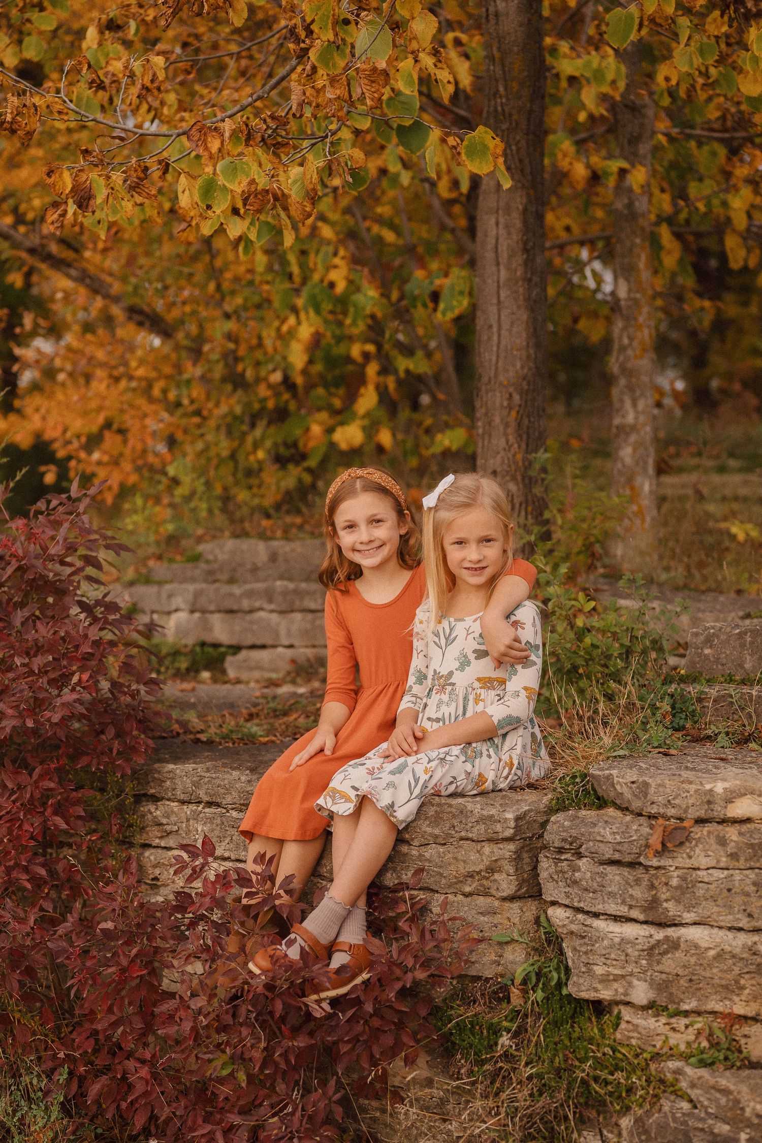 Two children sitting together on stone steps surrounded by vibrant fall foliage.
