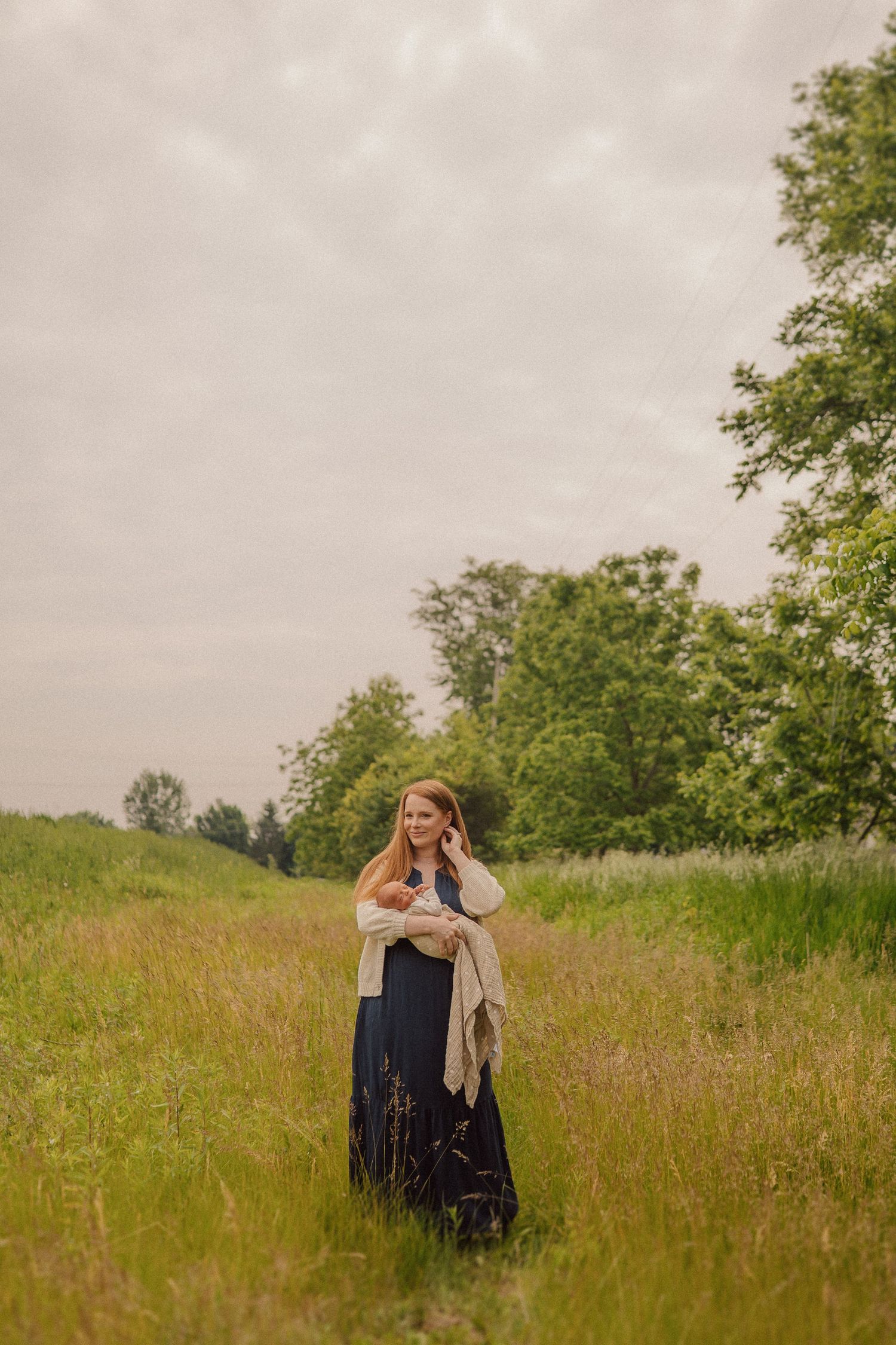 Person in long dark dress and shawl stands thoughtfully in a serene summer meadow surrounded by tall grass and trees.