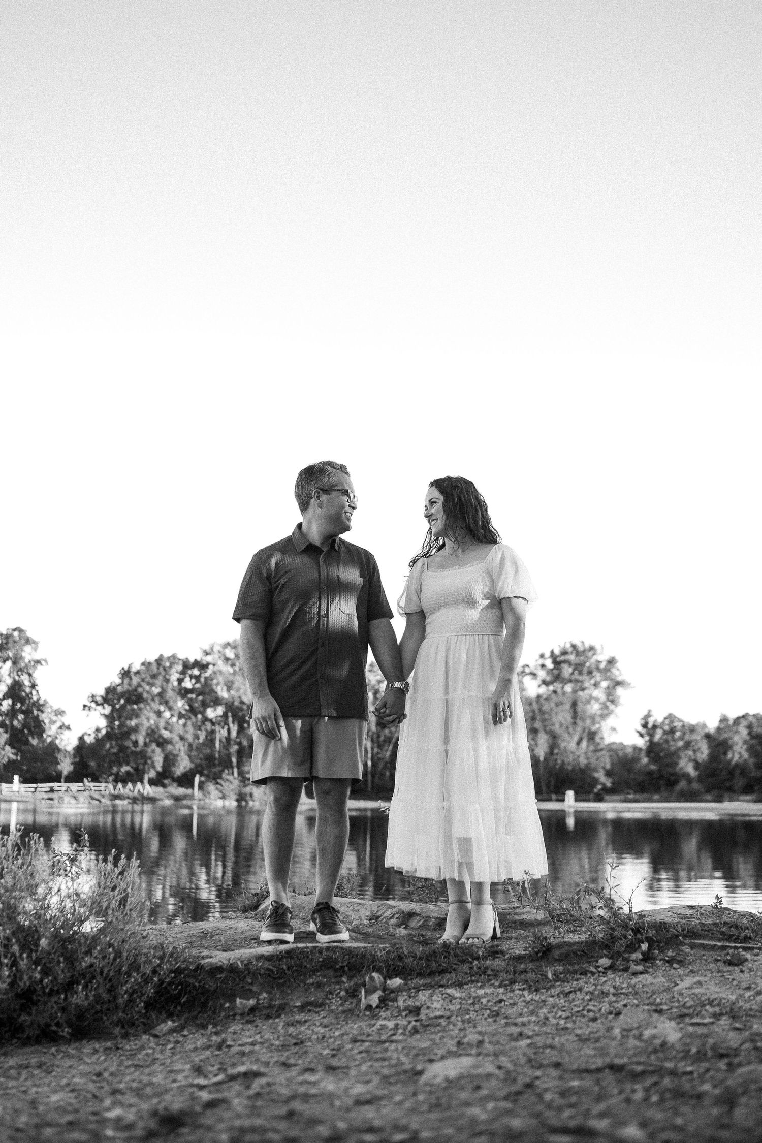 A couple in formal attire poses together near a lake at sunset creating silhouettes against the water.