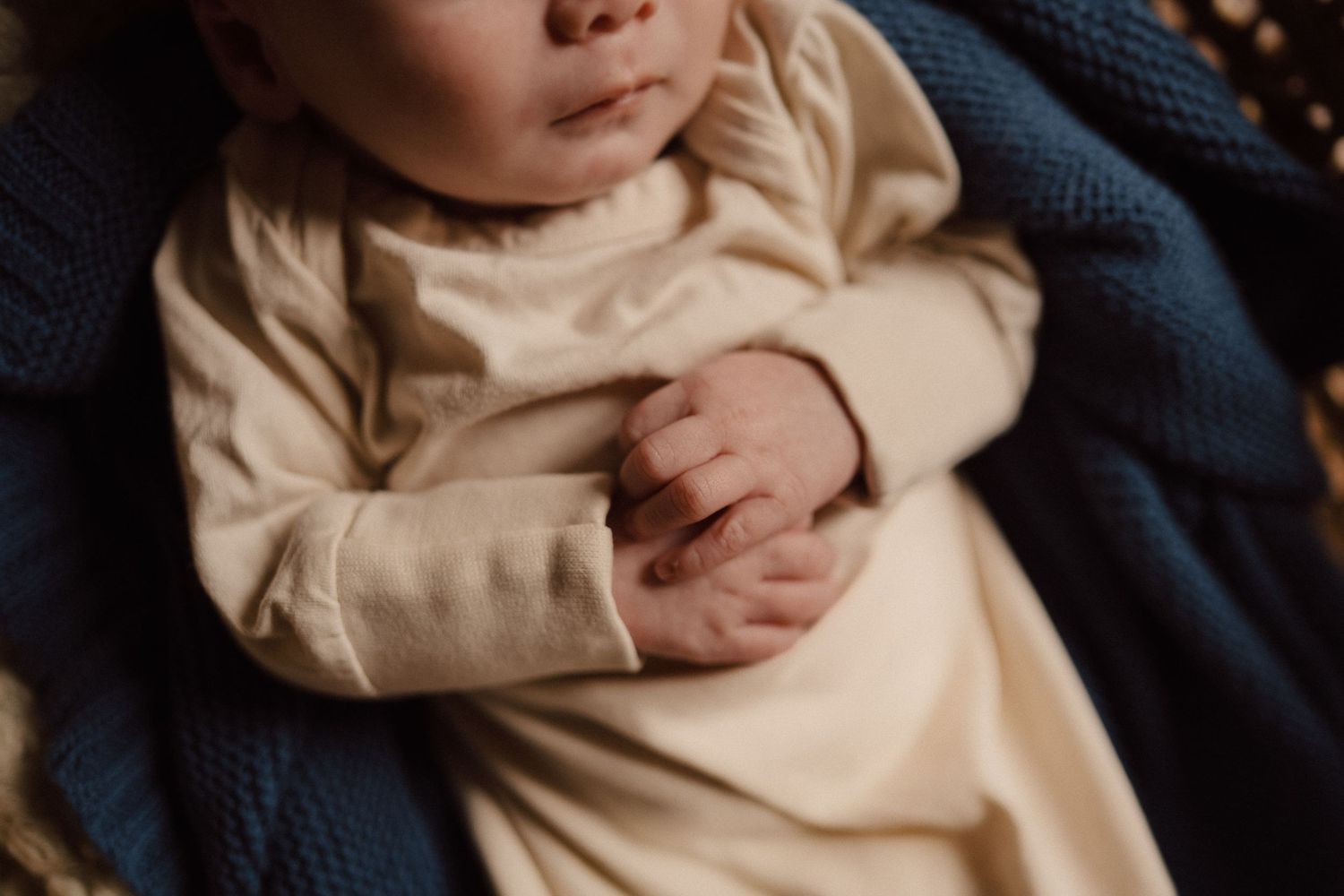 Close-up of tiny infant hands in cream colored clothing against blue fabric.