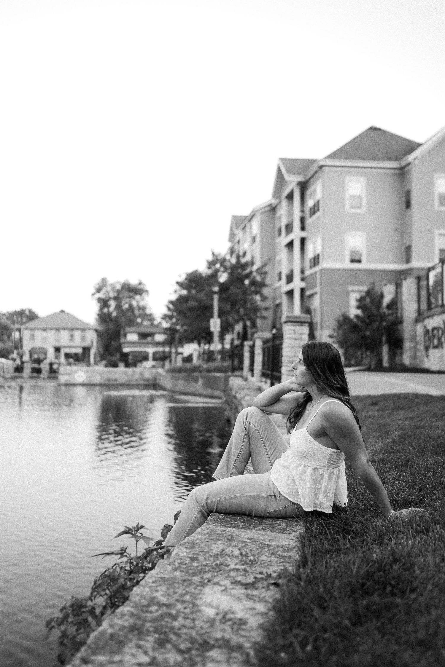 Person in white dress lounges by lakeside with residential buildings in background.