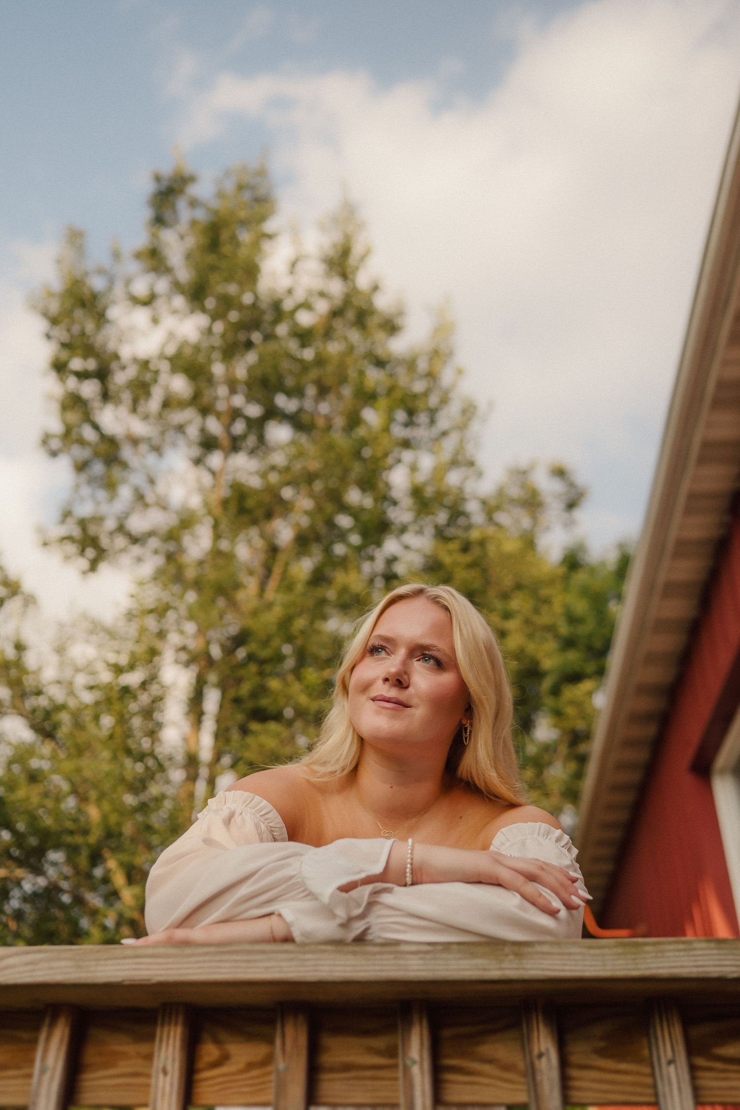 A person in a white top leans on a wooden deck railing with trees in the background.