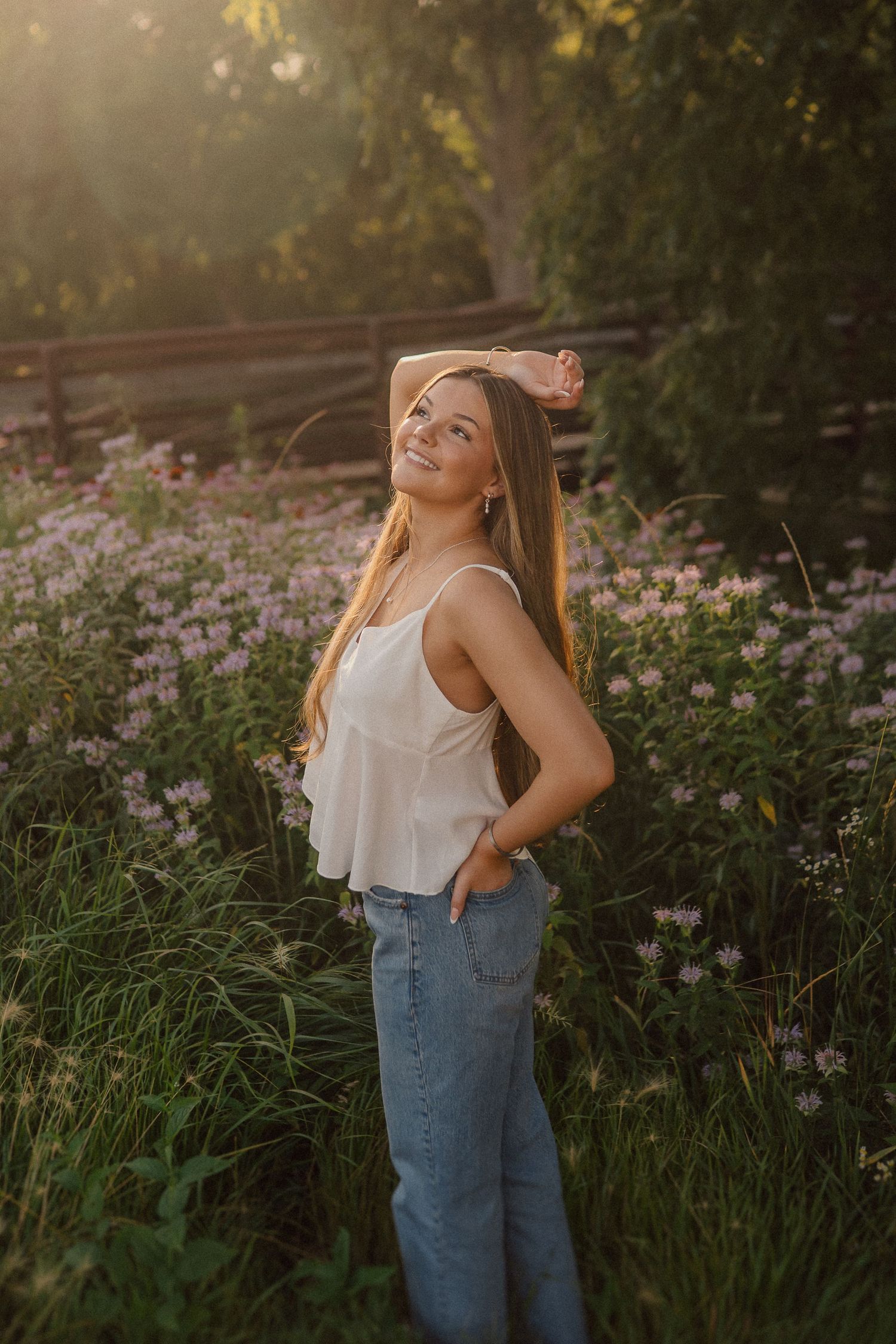 Person in white top and jeans poses among wildflowers during golden hour sunset.