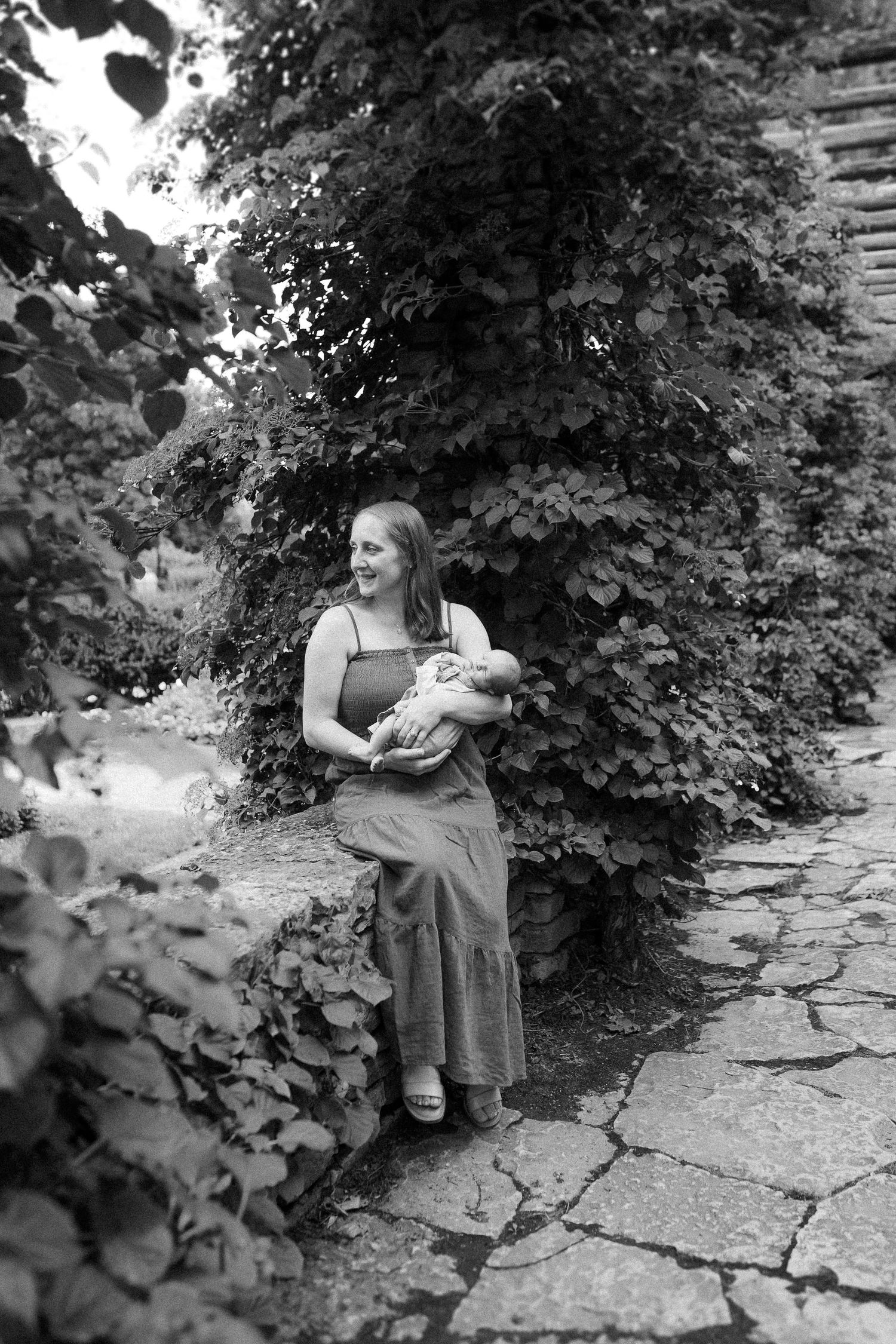 Black and white photo showing a person holding a baby while standing on a stone path surrounded by lush foliage.