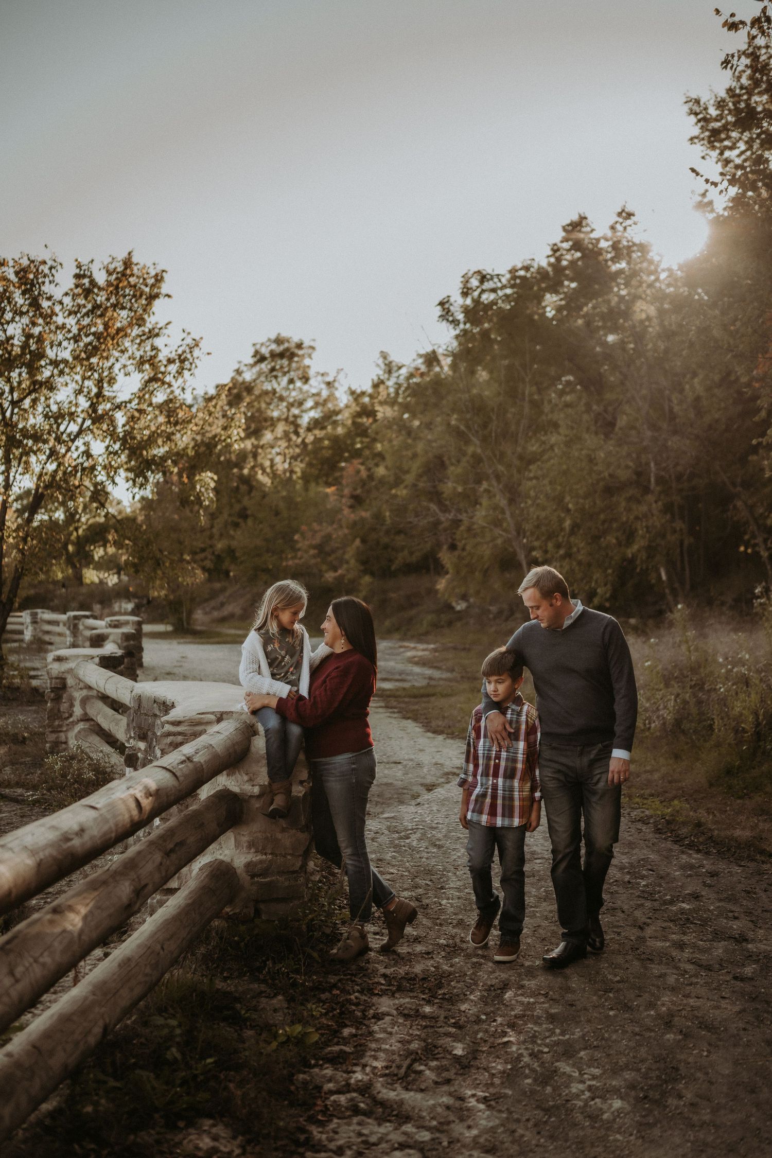 A family walks along a rustic dirt path with wooden fencing at sunset in an autumn forest setting.